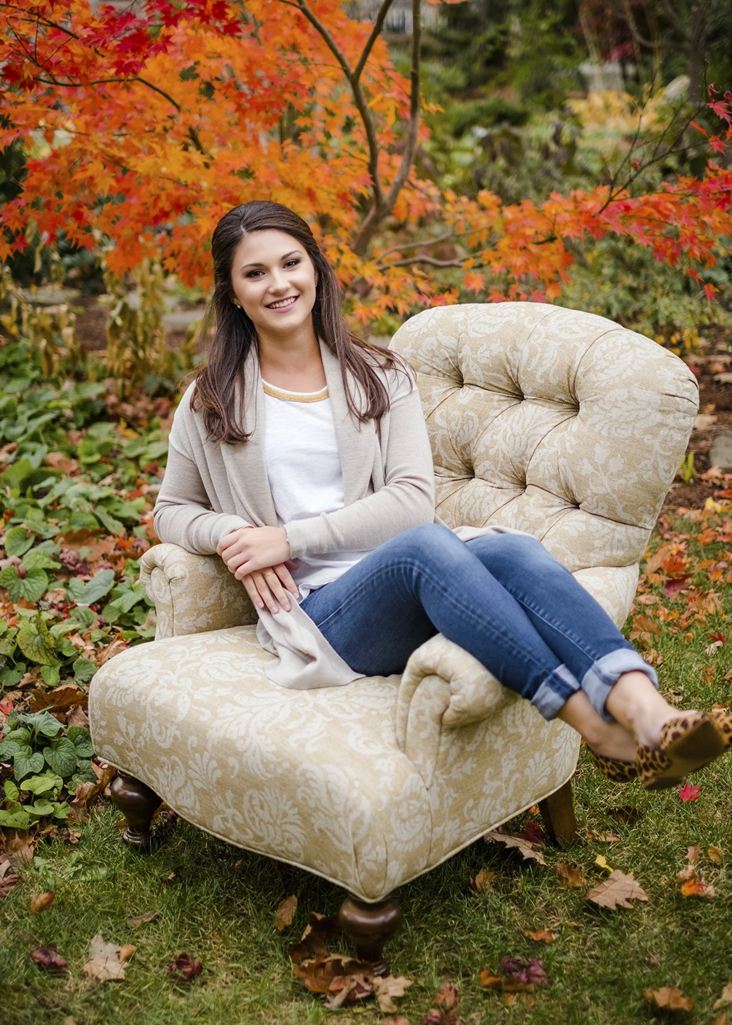 A smiling woman sitting on a cream-colored vintage armchair outdoors in a garden with colorful fall foliage in the background.