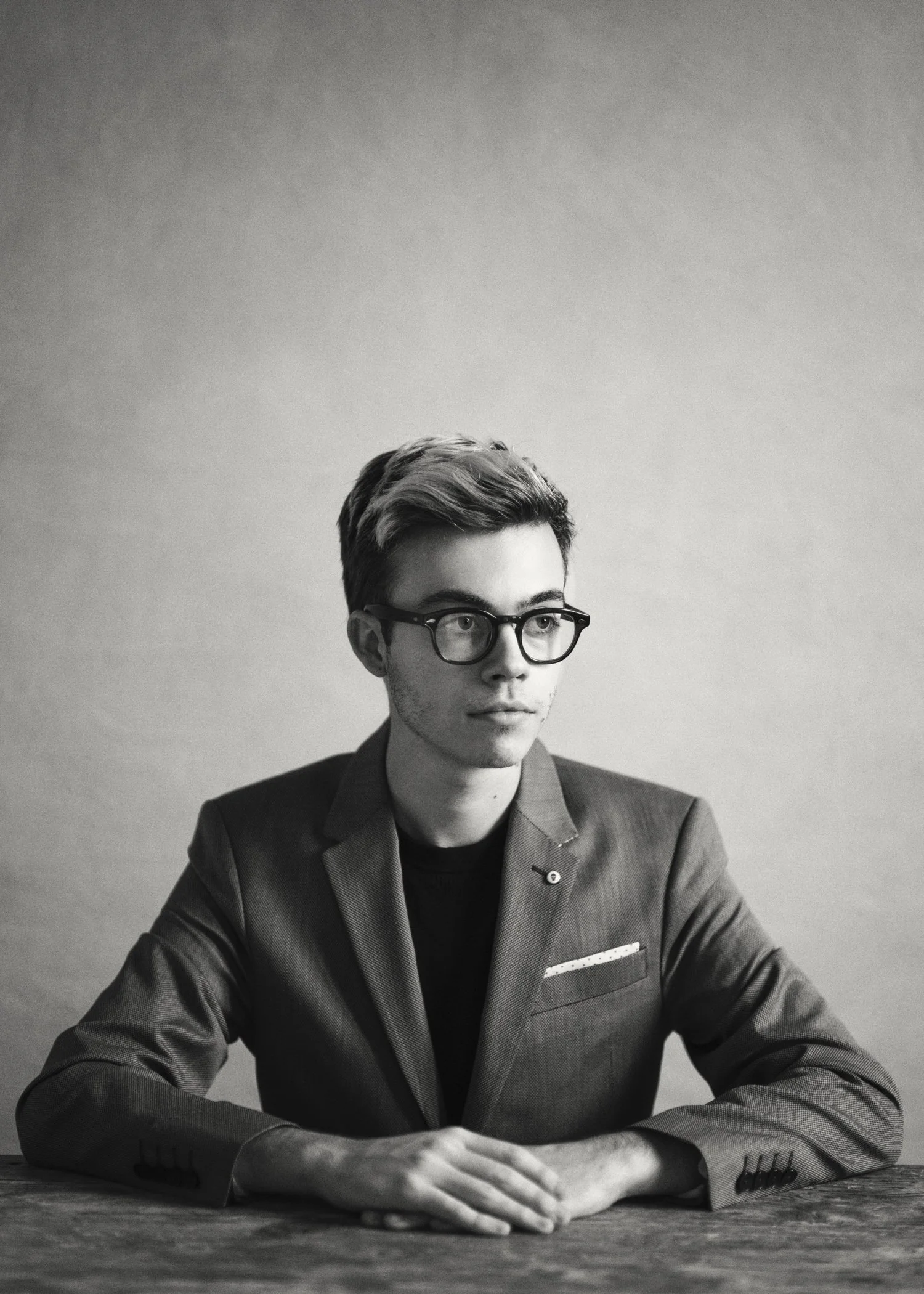 A young man with glasses and styled hair is sitting at a table, wearing a blazer over a dark shirt, looking off to the side in a black-and-white photo.