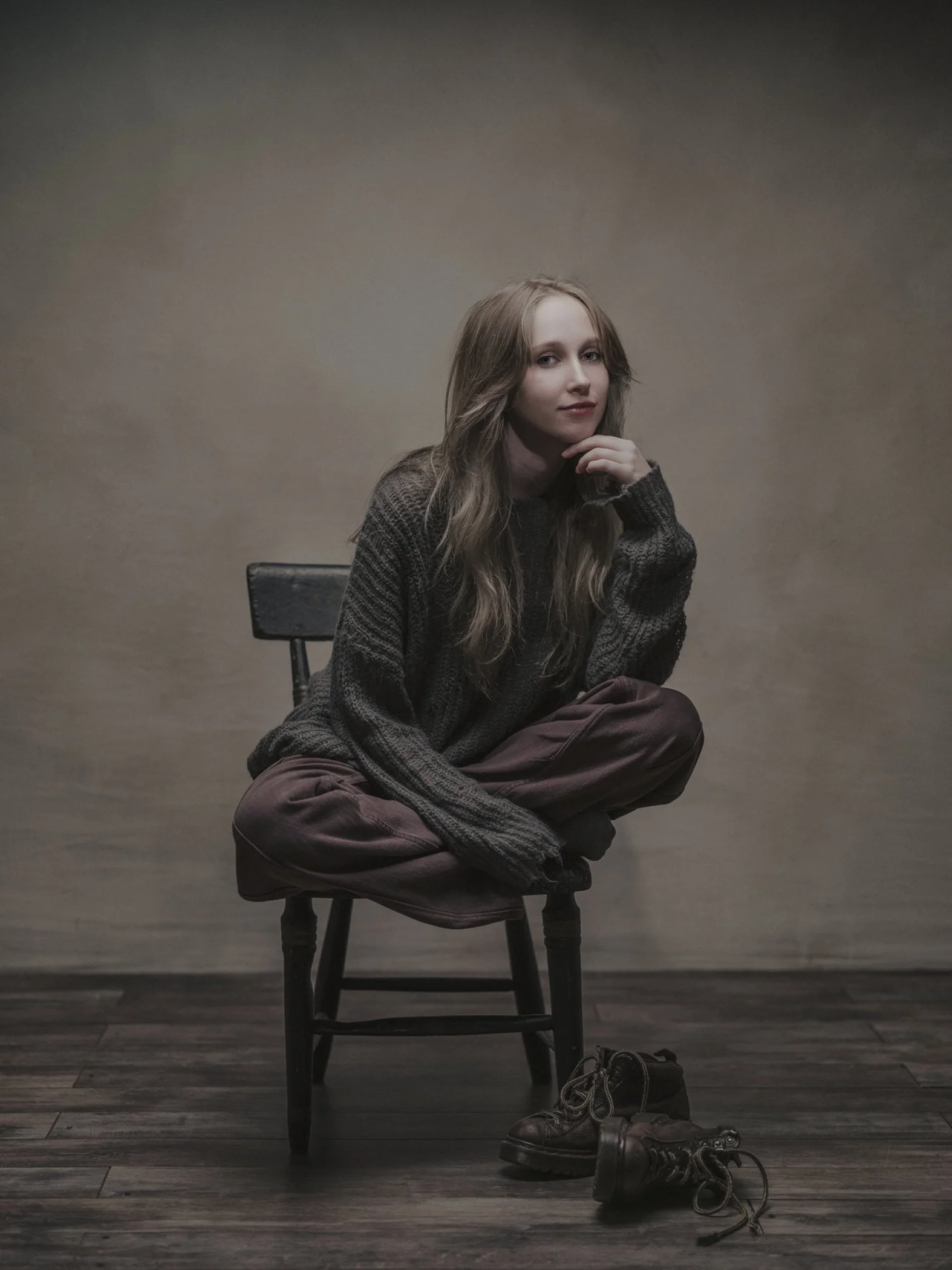 A young woman with long, wavy hair sitting on a wooden chair against a neutral-toned wall. She is wearing a dark sweater and brown pants, with her right hand resting on her chin and a subtle smile on her face. There are shoes and shoelaces on the flo