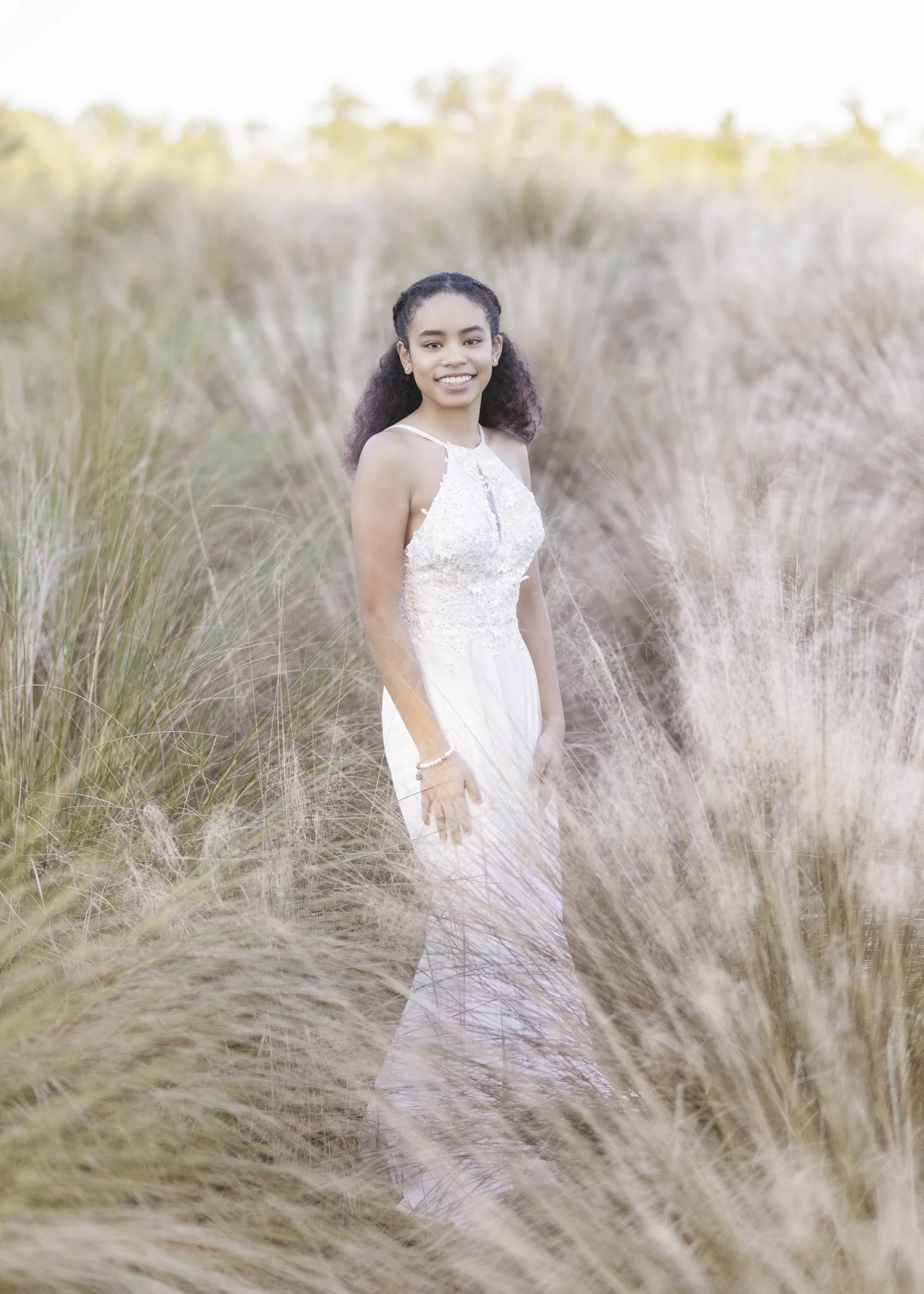 A young woman in a white dress standing in a field of tall, dry grass with blurred trees in the background.