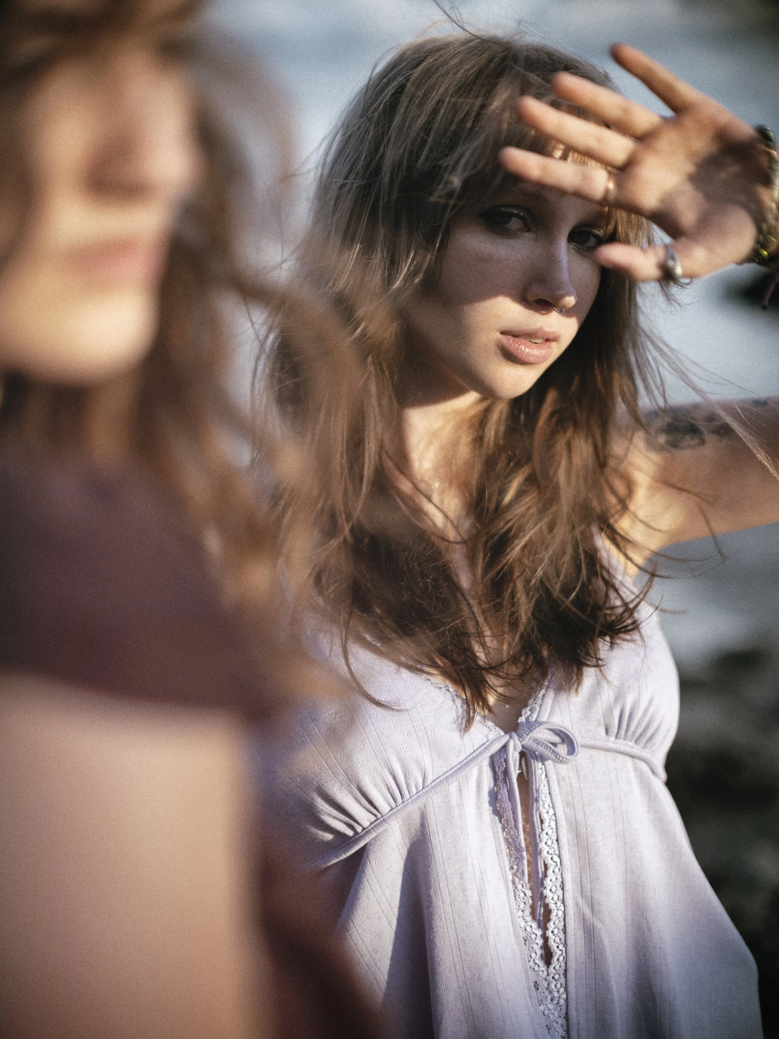 A woman with long wavy hair shielding her eyes from the sun, wearing a white blouse with lace details.