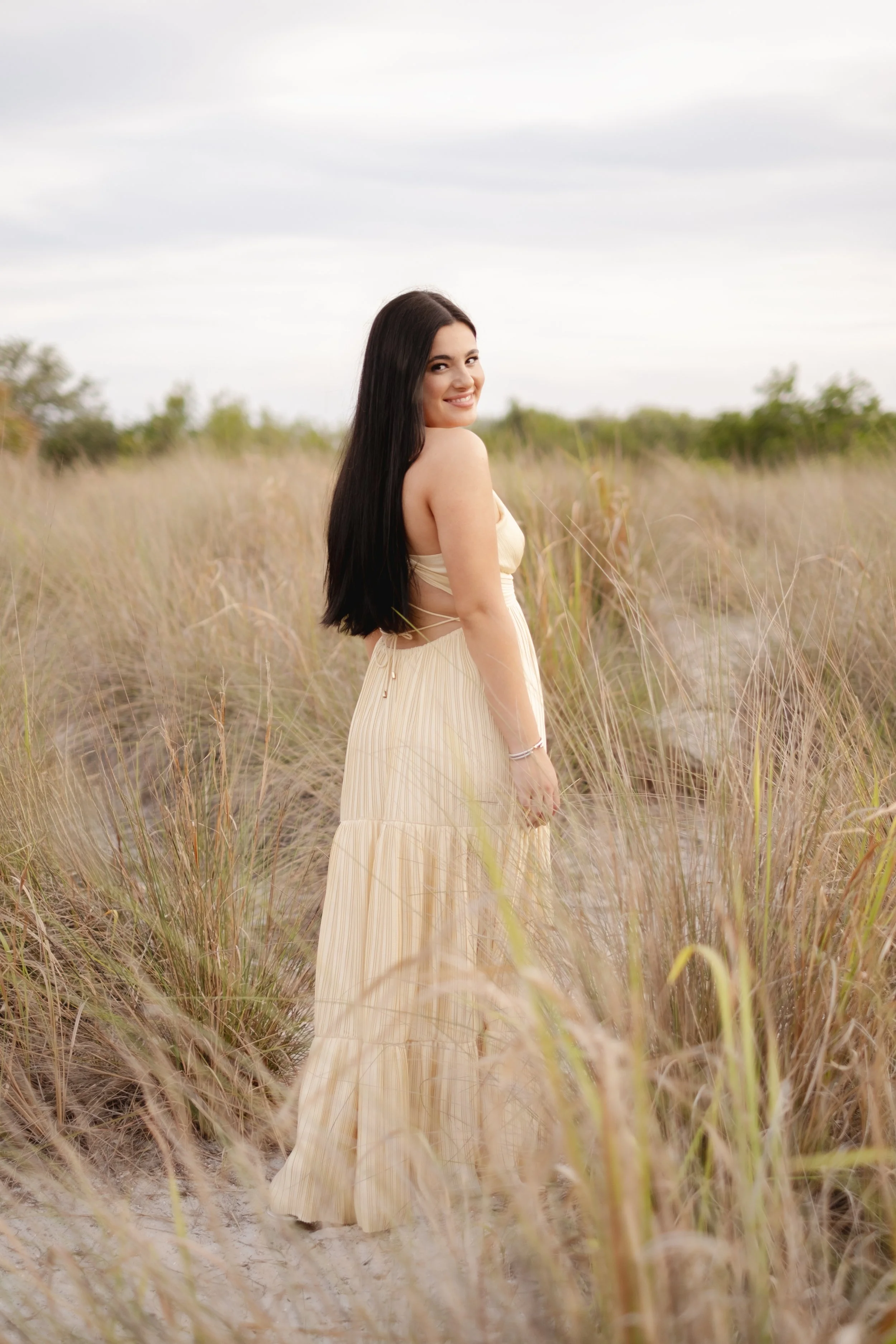 A woman with long dark hair in a light-colored, strapless dress standing in a sandy, grassy field with a cloudy sky in the background.