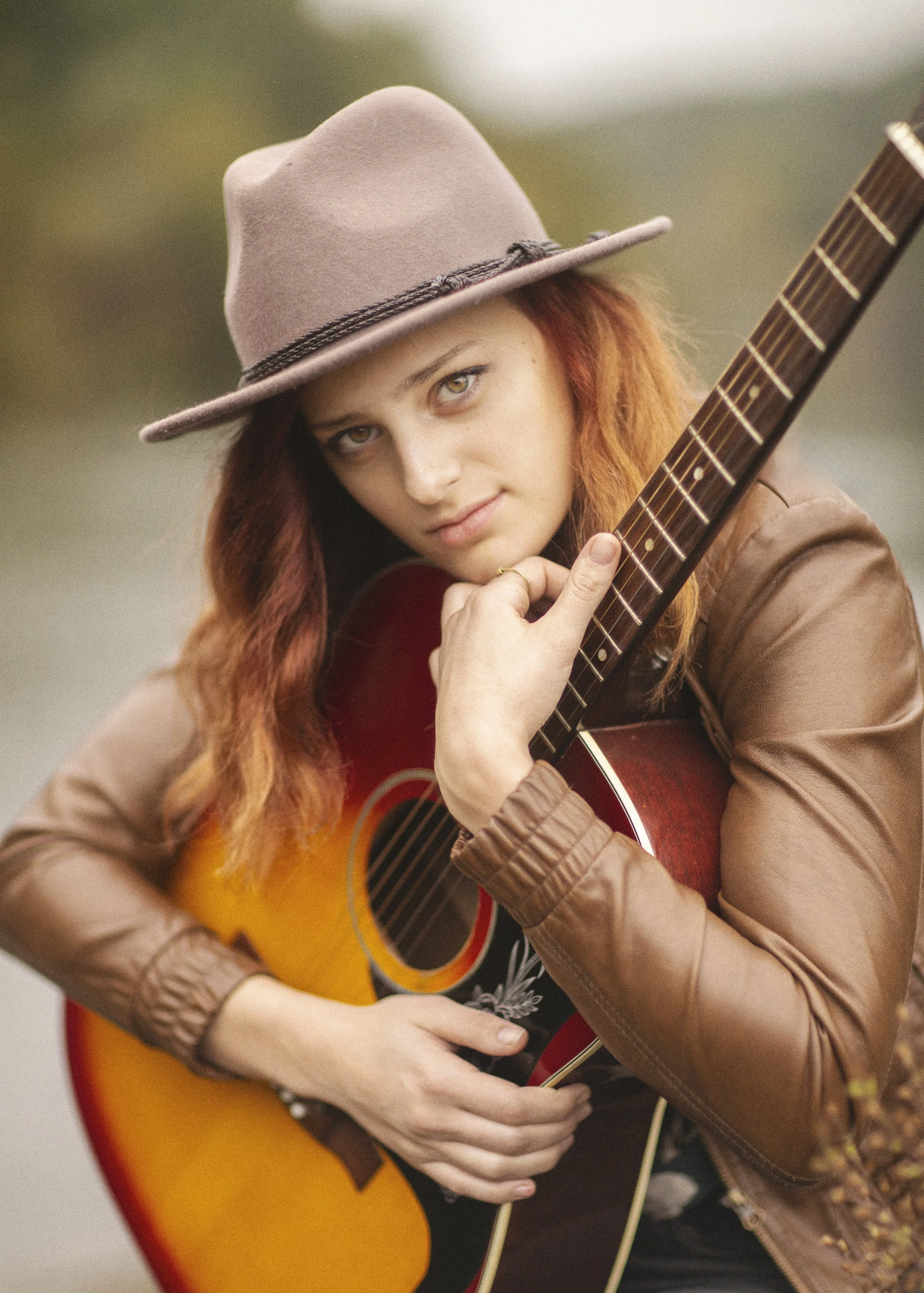 A young woman with long red hair, wearing a beige hat with a black band, holding an acoustic guitar and looking directly at the camera.