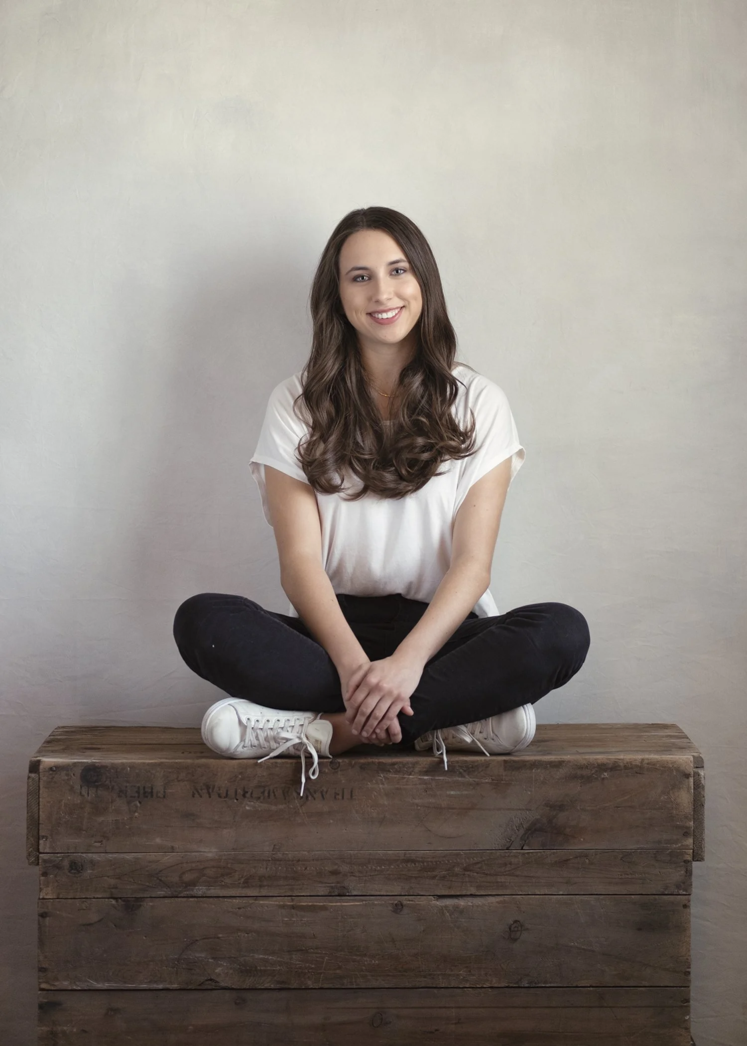 A young woman with long brown hair, wearing a white t-shirt and black pants, sitting cross-legged on a wooden crate in front of a plain white wall, smiling at the camera.