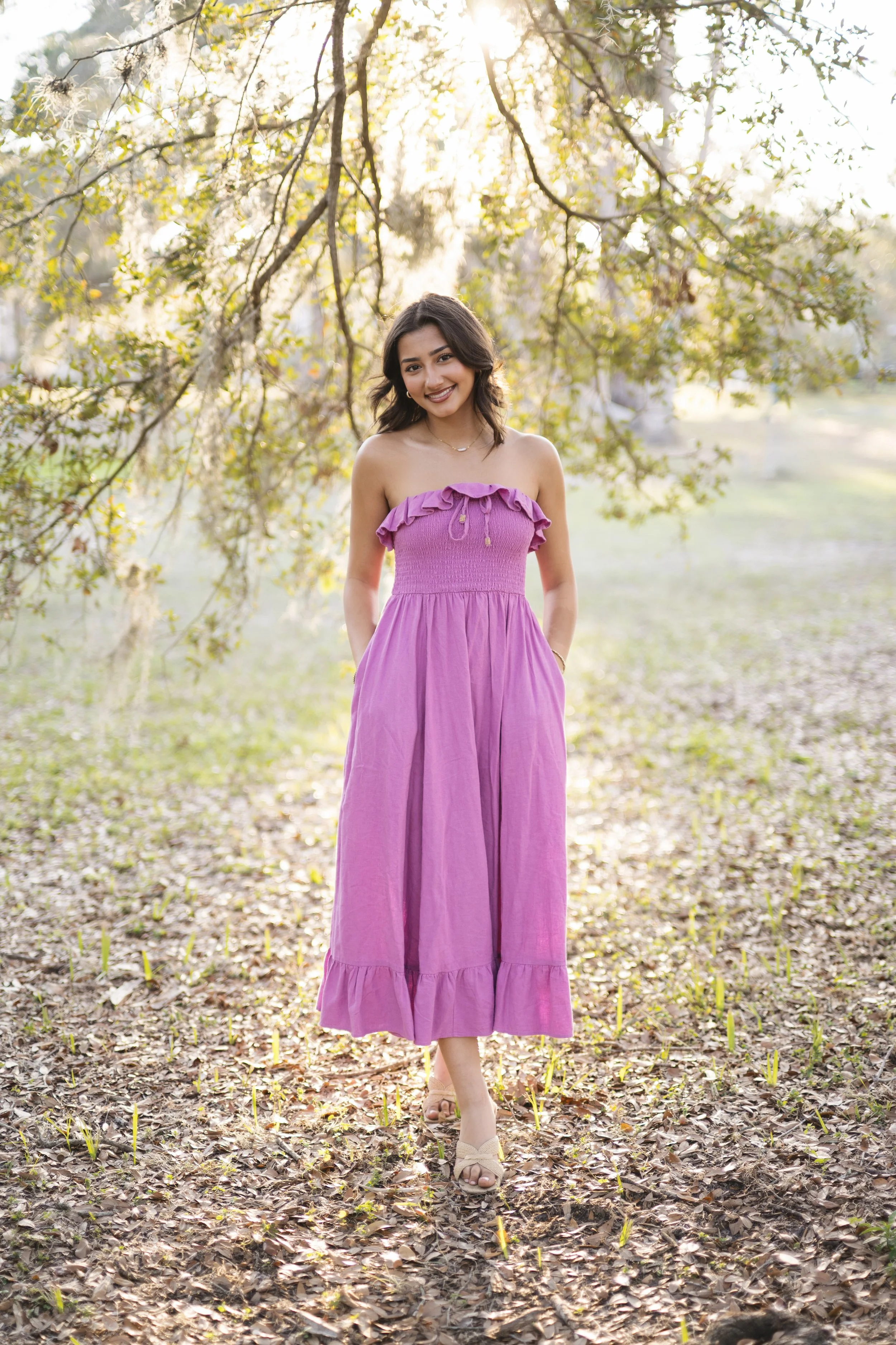 A young woman in a long purple dress standing outdoors on a path covered with fallen leaves, surrounded by trees with sunlight filtering through the branches.