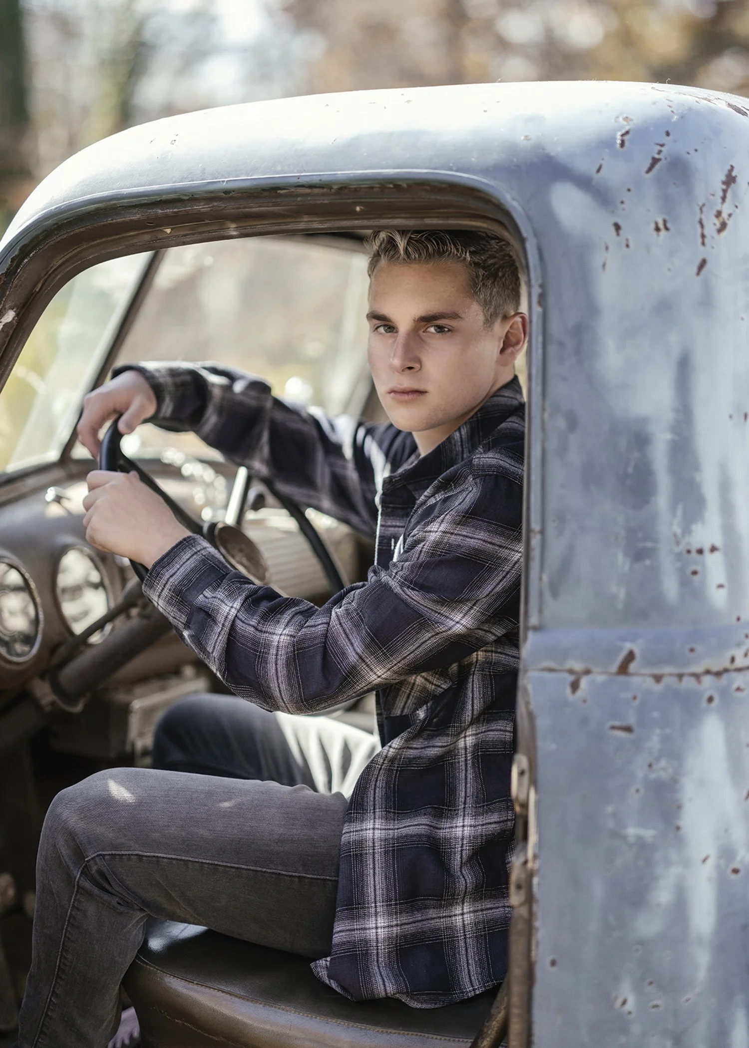 A young man with blonde hair and a serious expression sits in the driver's seat of an old, weathered pickup truck. He is wearing a black and grey plaid shirt and grey pants, with one arm resting on the steering wheel. The truck's interior appears age