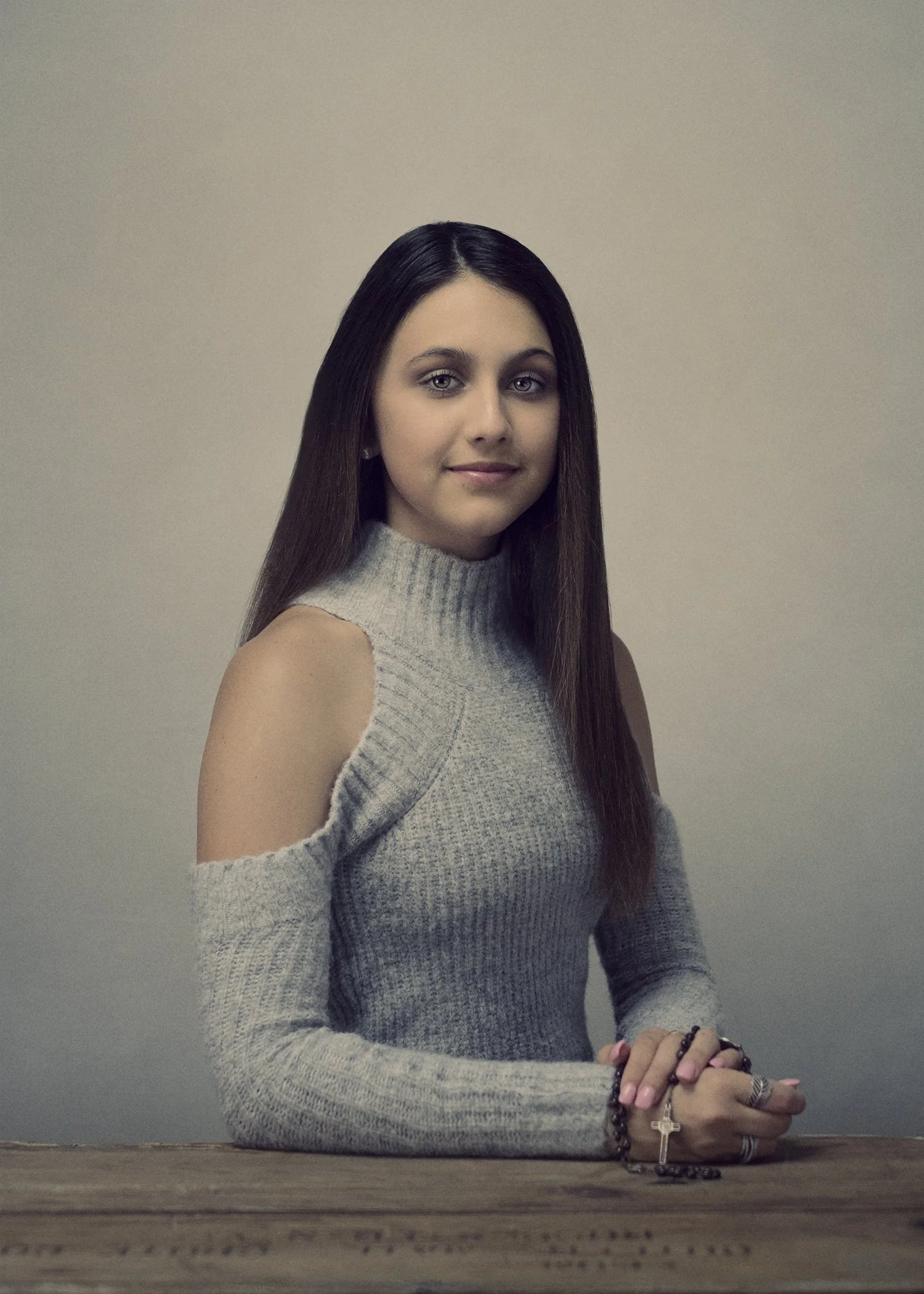 A young woman with long dark hair, wearing a gray sleeveless turtleneck sweater, is sitting at a wooden table against a plain wall. She is holding rosary beads with a cross in her hands, which are resting on the table, and is looking at the camera wi