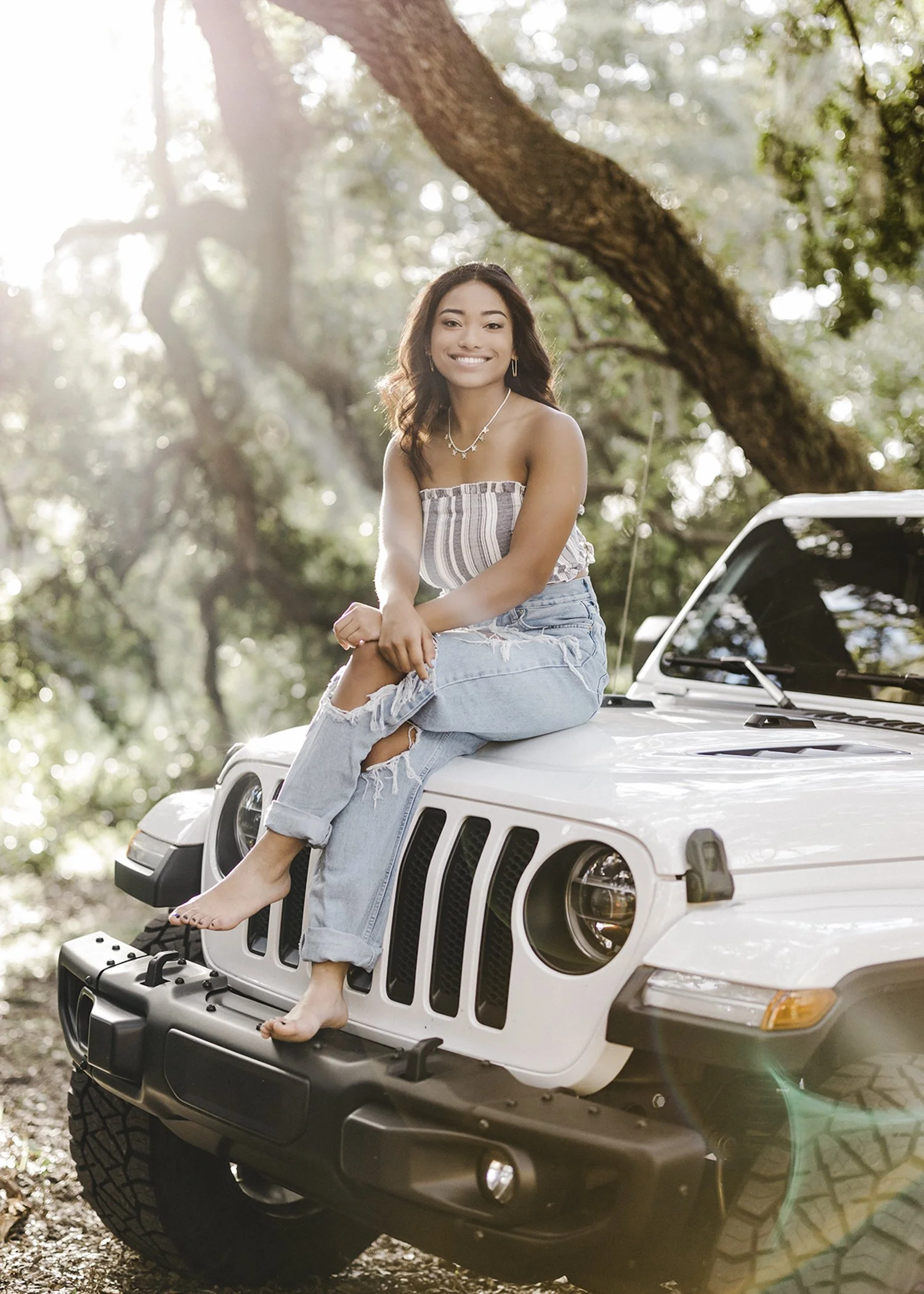 A young woman sitting on the hood of a white Jeep vehicle outdoors surrounded by trees, smiling at the camera during daylight.