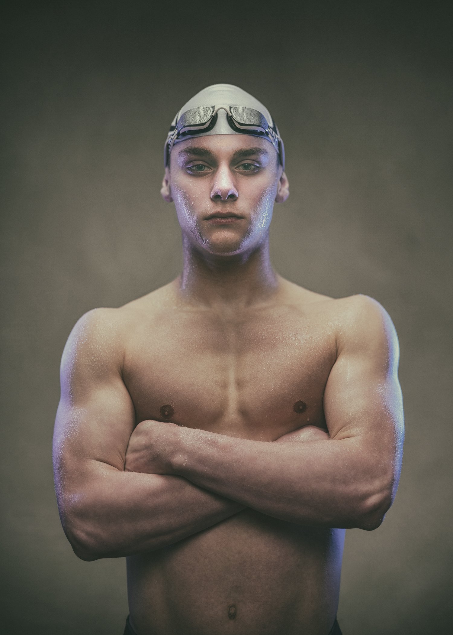 A young man with short hair and swimming goggles on his forehead, standing shirtless with arms crossed against a dark, gradient background.