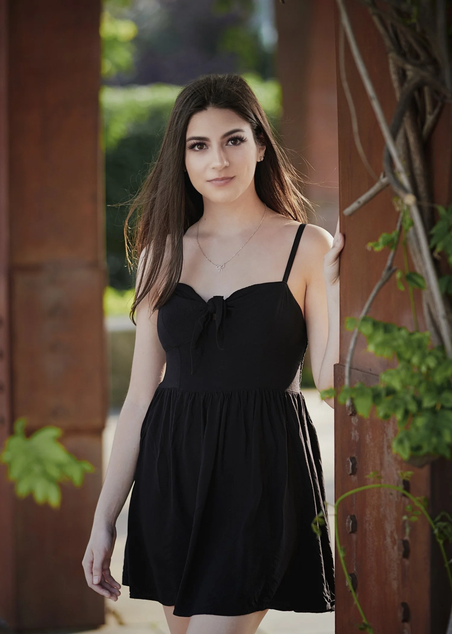 A young woman with long dark hair wearing a black dress with spaghetti straps, standing outdoors beside a wooden structure with greenery behind her.