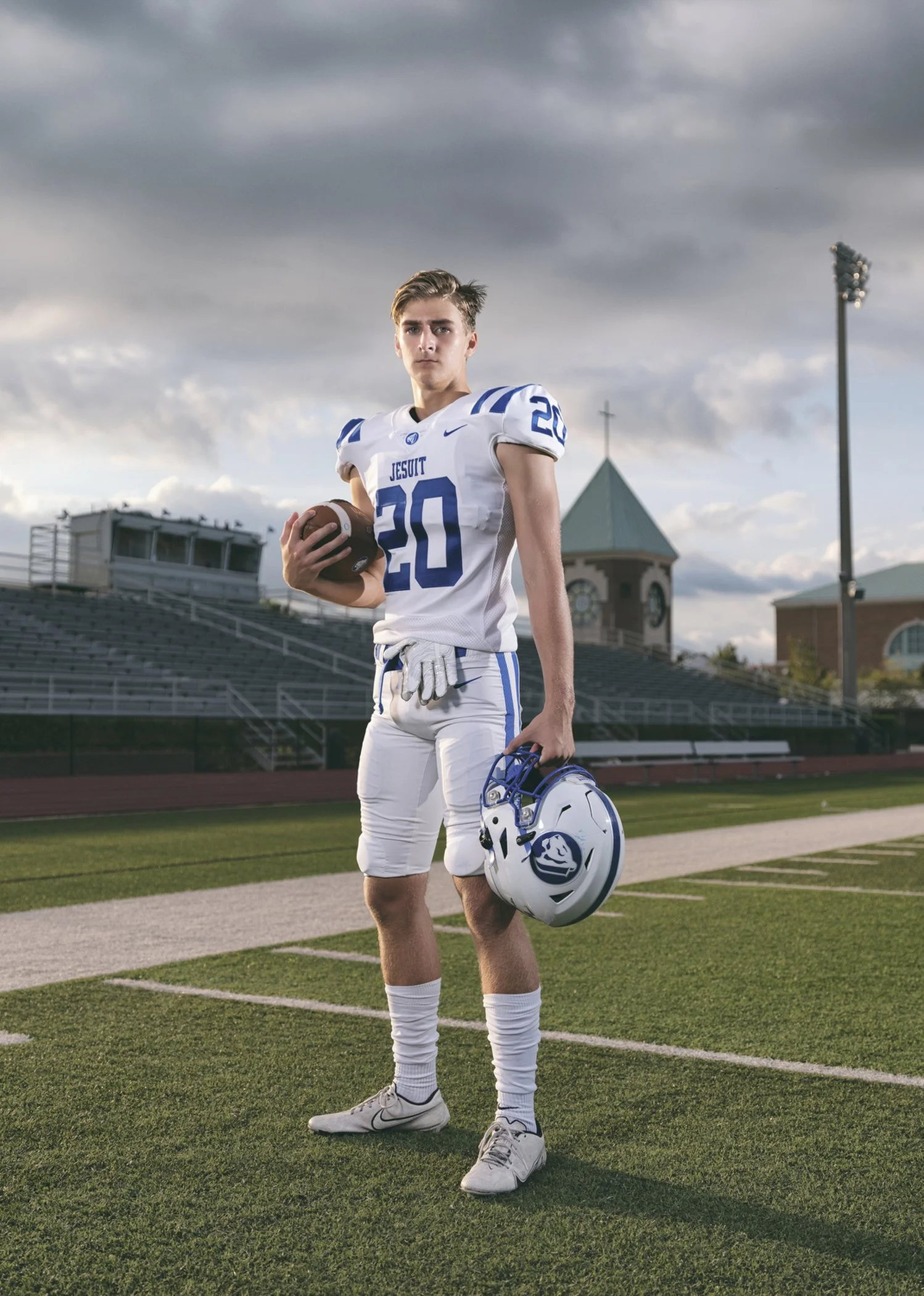 A young male football player stands on a football field, holding a football in one hand and a helmet in the other. He is dressed in a white and blue uniform with the number 20, under a cloudy sky.