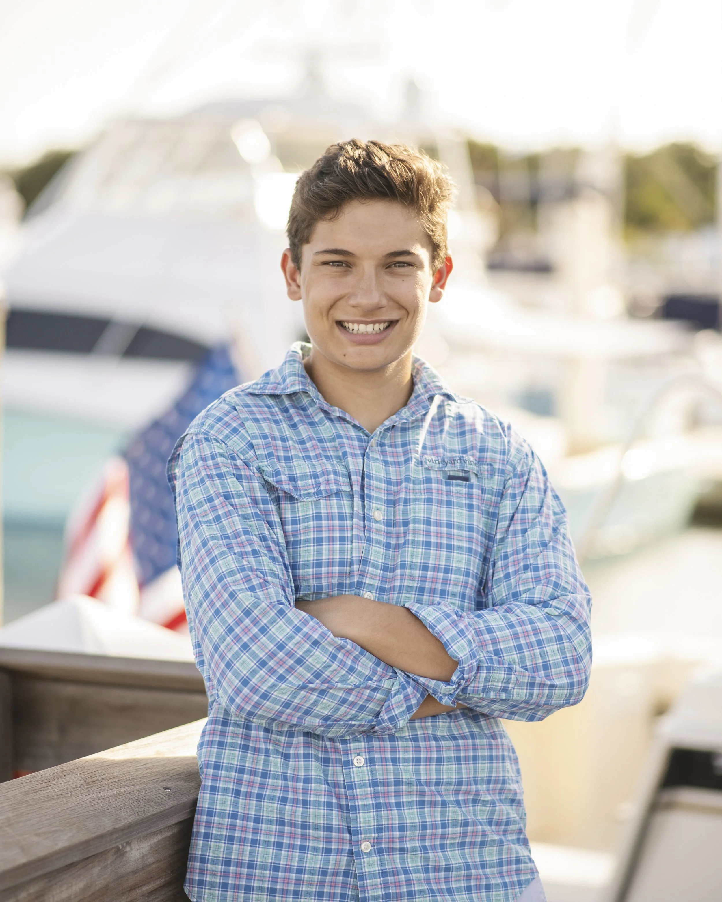 A young man with short brown hair and a checkered shirt smiling at the camera, standing outdoors near boats and a wooden railing.