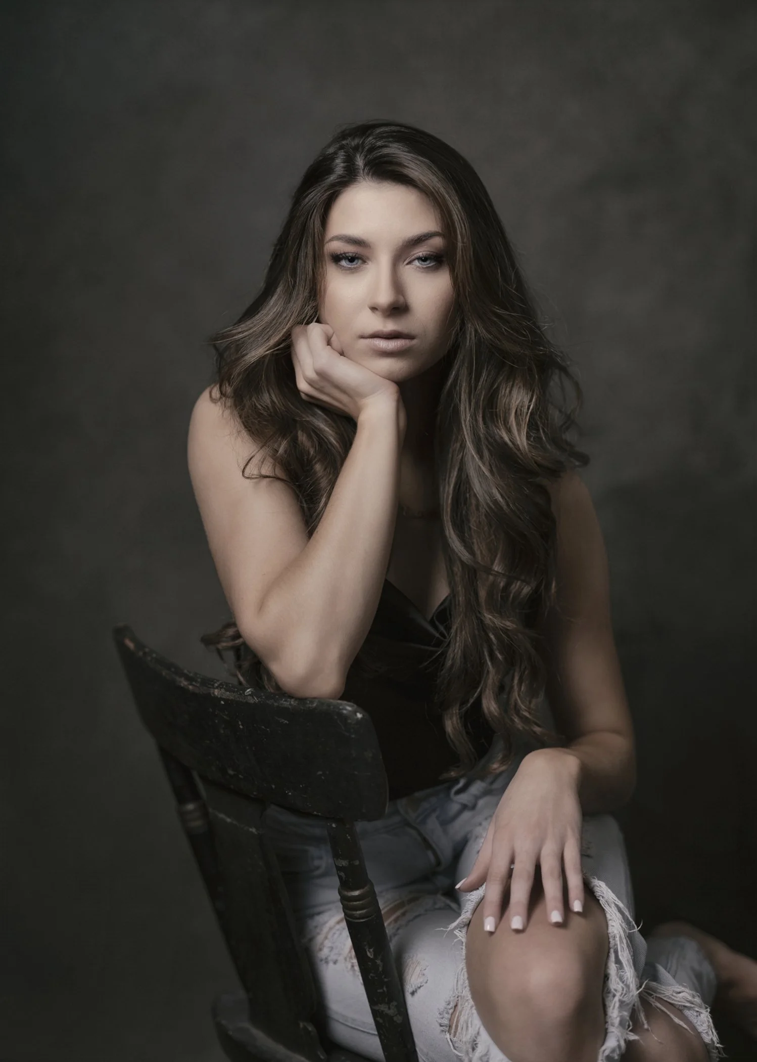 A young woman with long, wavy brown hair resting her chin on her hand, seated on a black vintage chair with a dark, textured background.