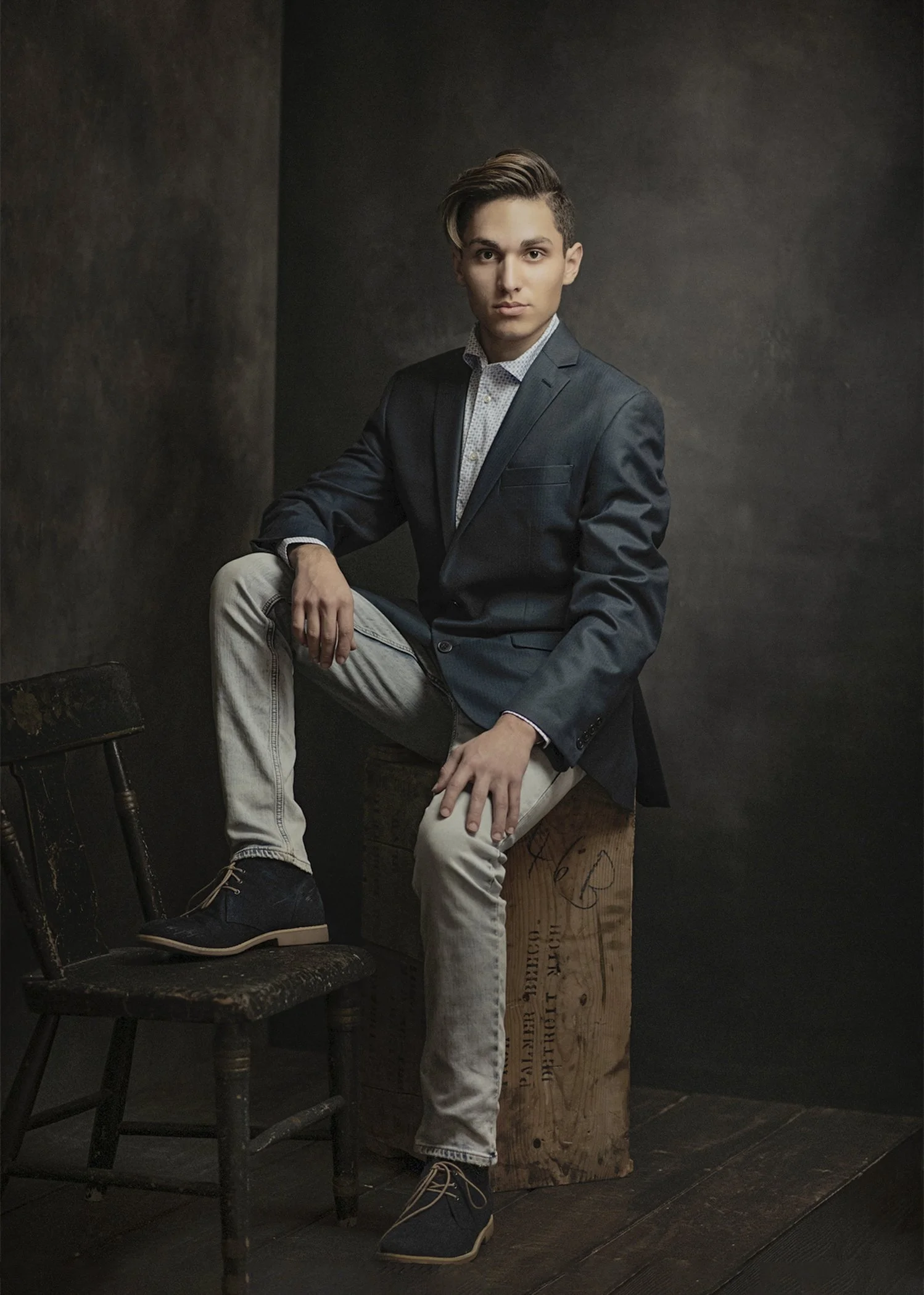 Young man in a dark suit and casual light-colored pants sitting on an old wooden chair, posed against a dark background with a wooden box behind him.