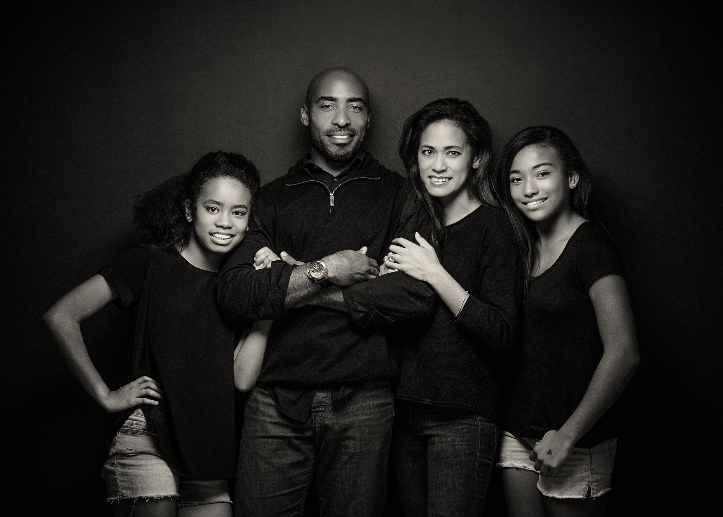 Group of five people, three women and two men, standing close together and smiling for a photo against a black background, in black and white.