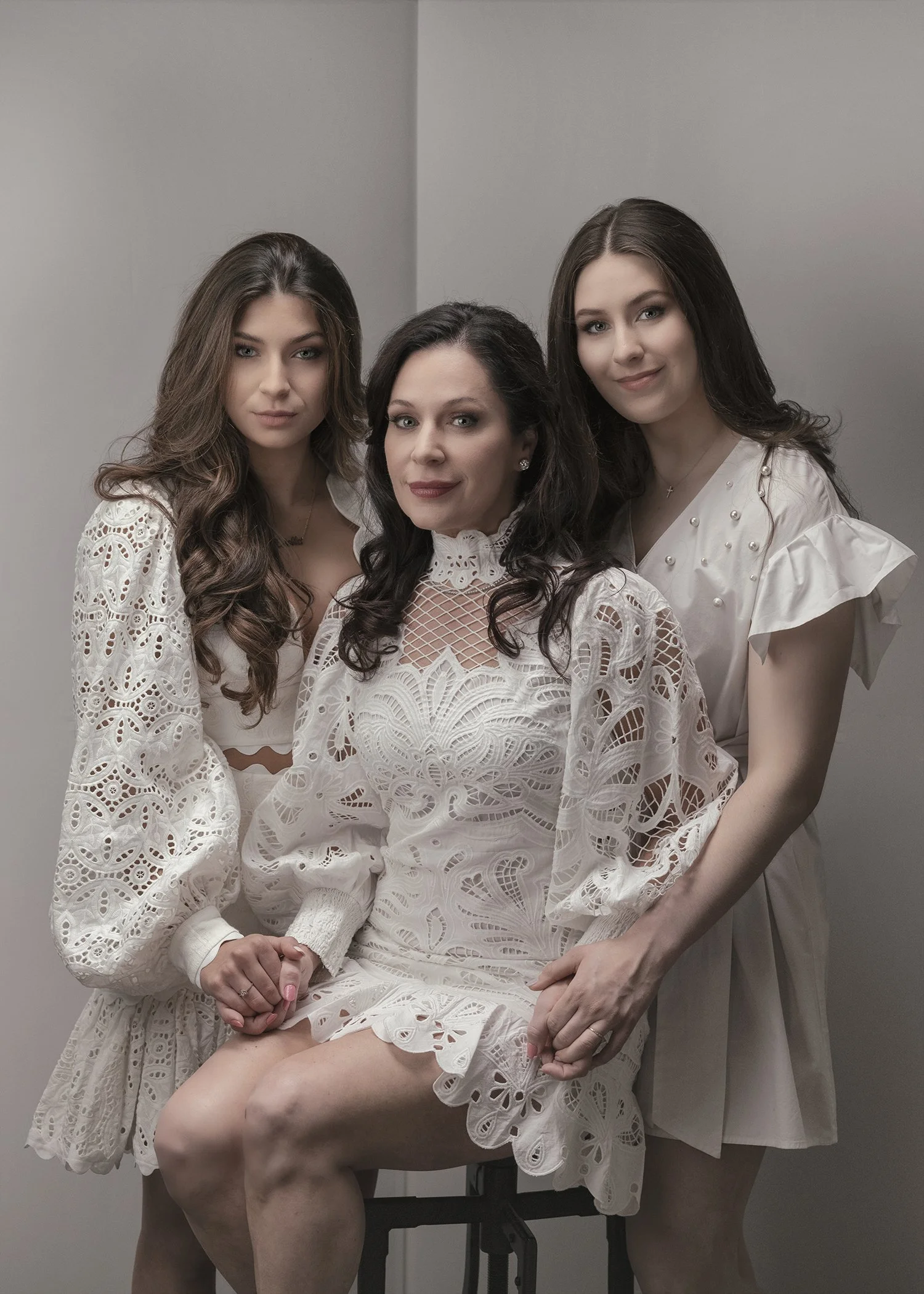 Three women dressed in white with lace and eyelet patterns posing against a plain gray wall. The woman in the middle is seated with her hand on her knee, smiling, and the two women on either side are standing close, also smiling gently.