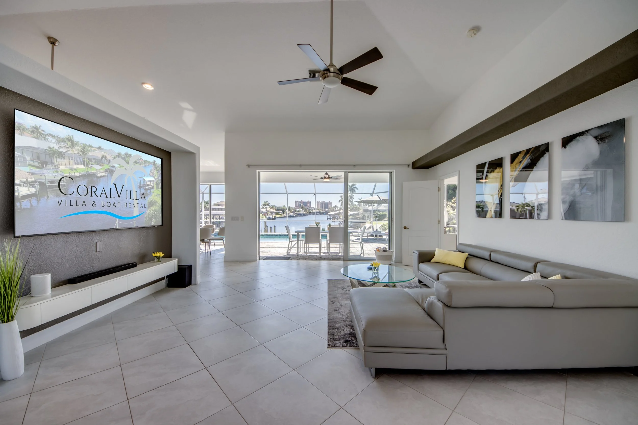 Bright living room with white tile floors, gray walls, white sectional sofa, glass coffee table with small yellow flowers, large wall-mounted TV displaying 'Coral Villa Villa & Boat Rental,' sliding glass doors showing an outdoor patio, dock, and har