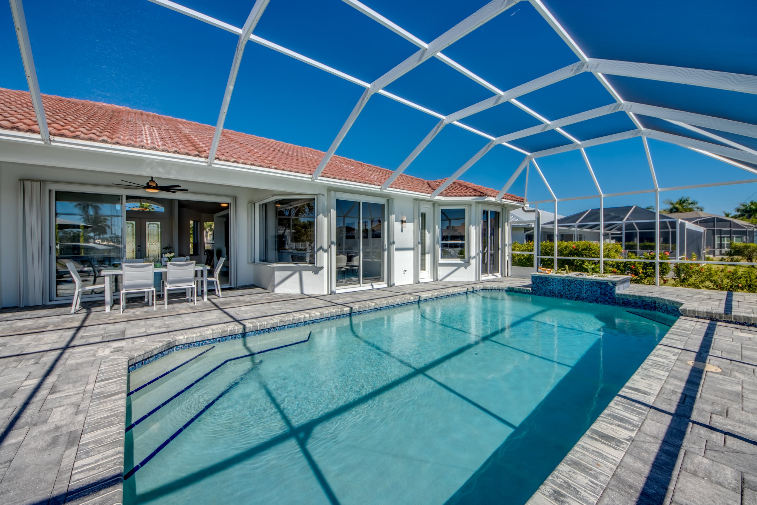 A backyard pool area with a swimming pool, patio furniture, and a house with a screened-in porch under a clear blue sky.