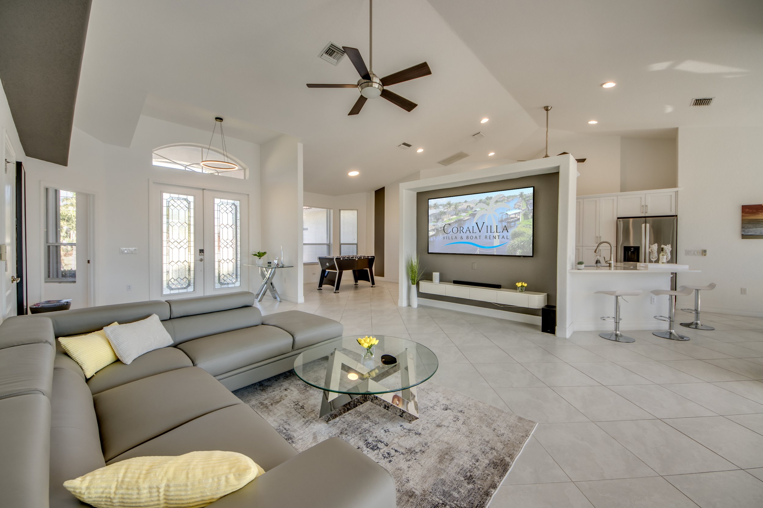 Modern living room with a large gray sectional sofa, glass coffee table, flat-screen TV, and a kitchen area with white cabinets and barstools.