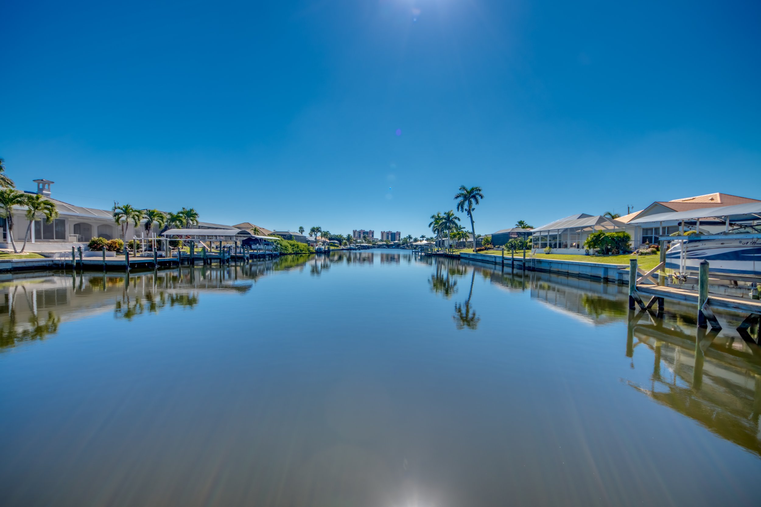 A canal lined with houses and palm trees, with boats docked along the water under a clear blue sky.