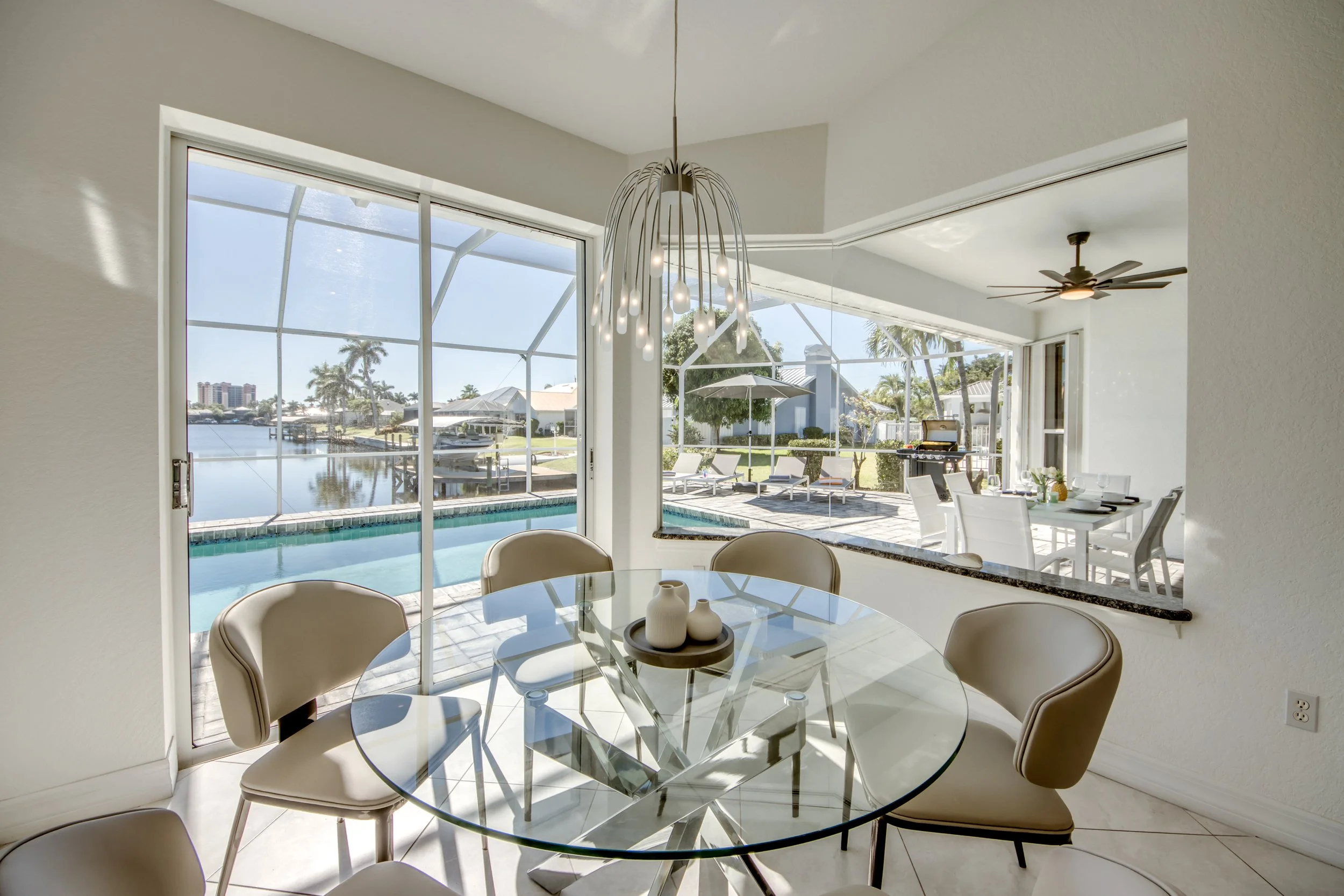 Interior view of a dining area with a glass table and six beige chairs, overlooking a screened pool area with outdoor furniture and a waterway in the background.
