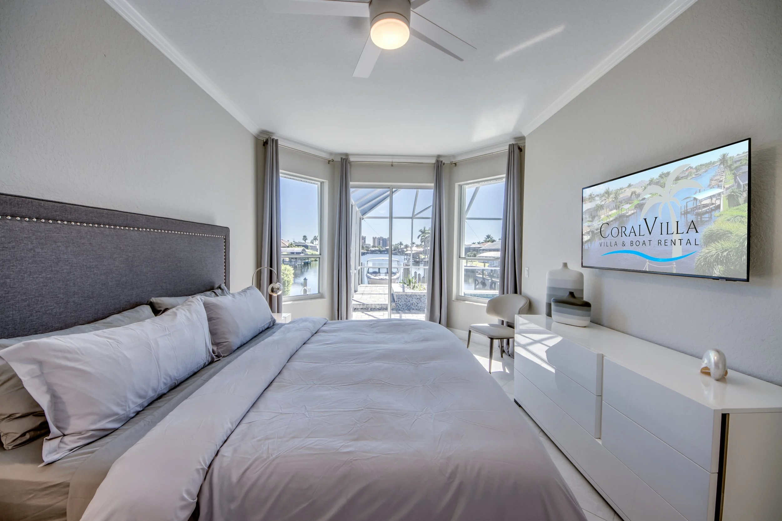 A bedroom with a large bed with gray headboard and white bedding, a white dresser, a TV displaying "Coral Villa Villa & Boat Rental," three windows with curtains showing a marina and water outside, and a ceiling fan.