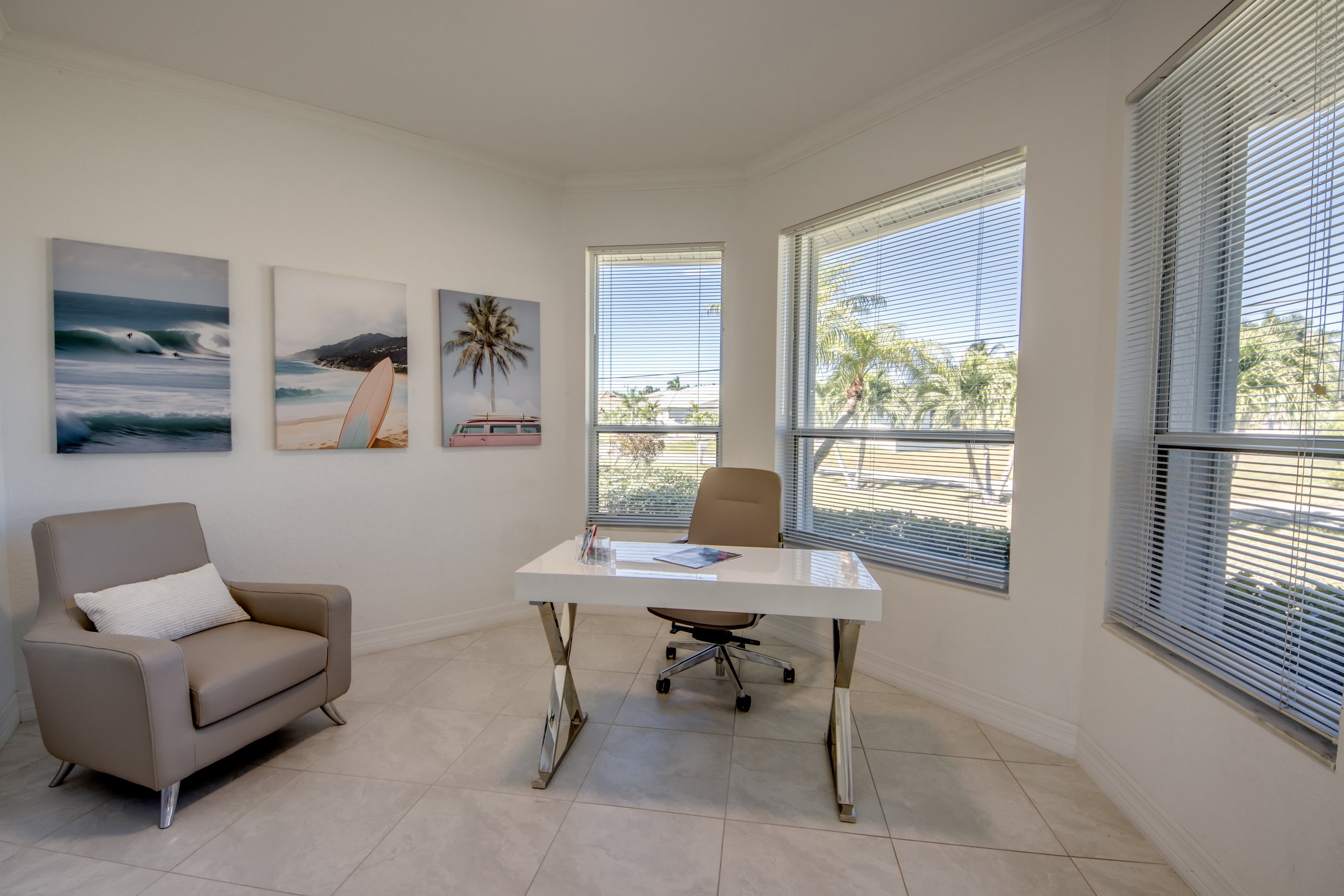 Office room with large windows, a white desk with a black chair, a beige armchair with a white pillow, and three framed beach-themed photographs on the wall.