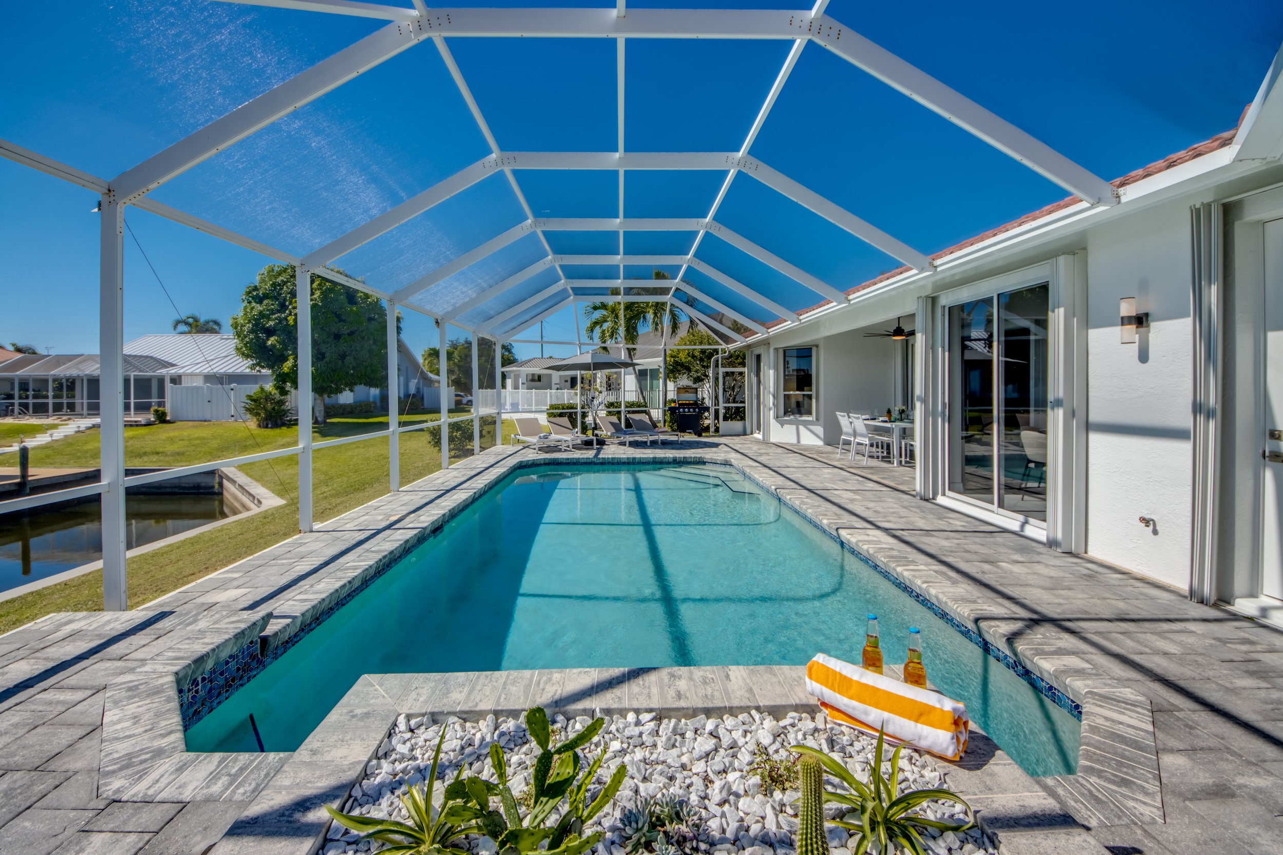Backyard swimming pool with a screened enclosure, outdoor furniture, and a grill, under a clear blue sky.