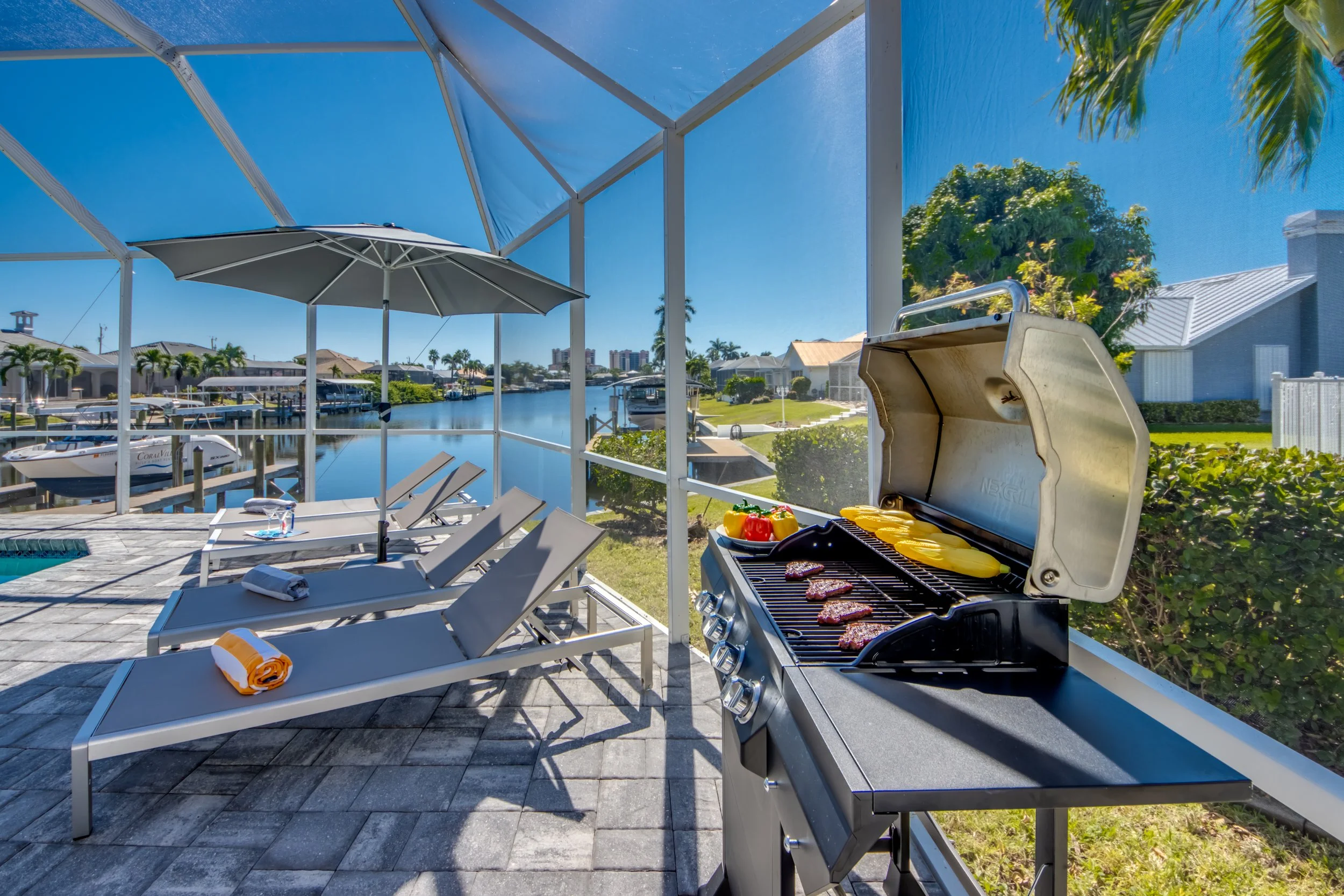 View of a backyard patio with lounge chairs, a grill with steaks, and a canal with boats, houses, and palm trees in the background under a clear blue sky.