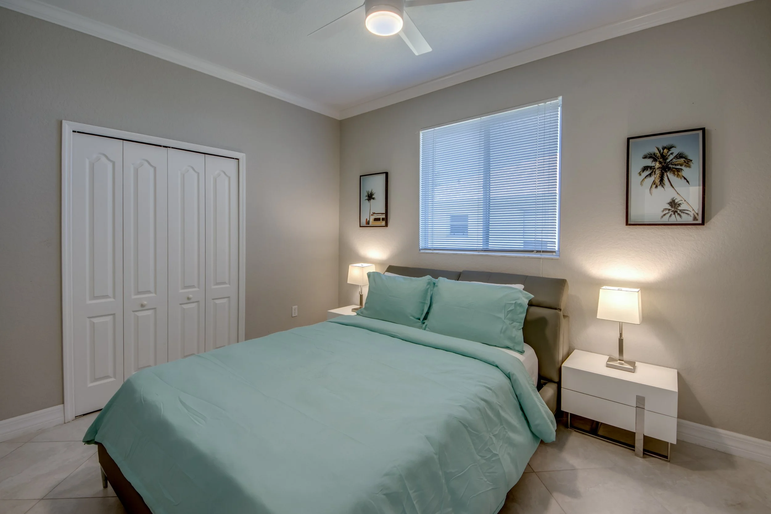 A bedroom with a bed covered in light green bedding, two nightstands with lamps, artwork of palm trees on the wall, a window with blinds, and a ceiling fan.