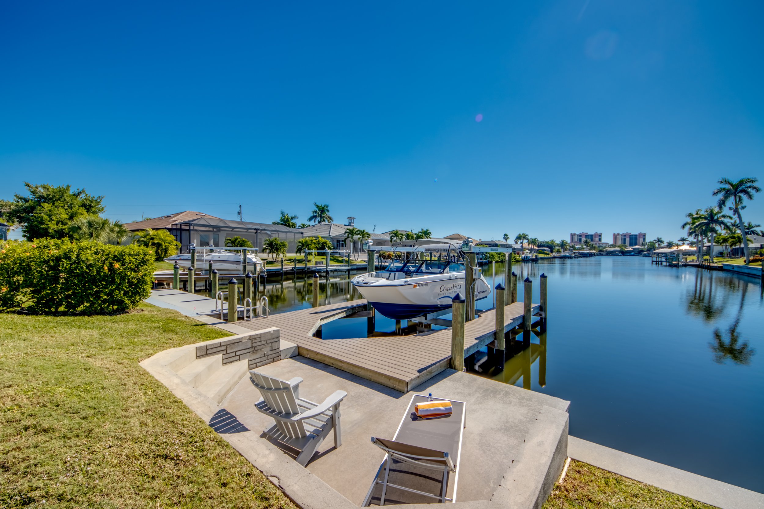 A cozy waterfront scene showcasing a dock with boat slips, small boats, and lounge chairs on a sunny day with clear blue sky, palm trees, and residential houses along the canal.