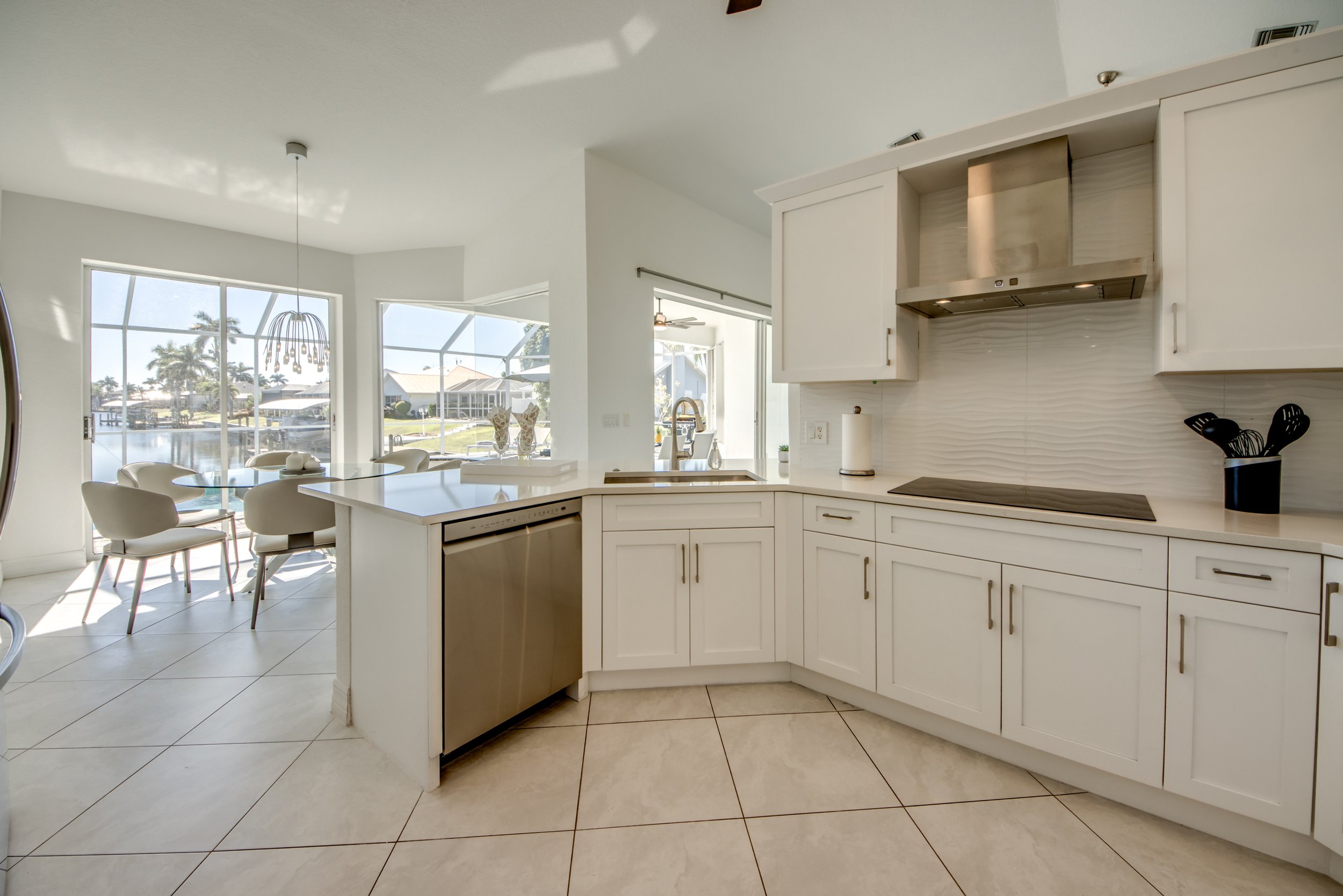 Modern kitchen with white cabinets, stainless steel range hood, and a tile backsplash. Adjacent to a dining area with a glass-top table and white chairs. Large windows and a screened-in porch overlooking a waterway.