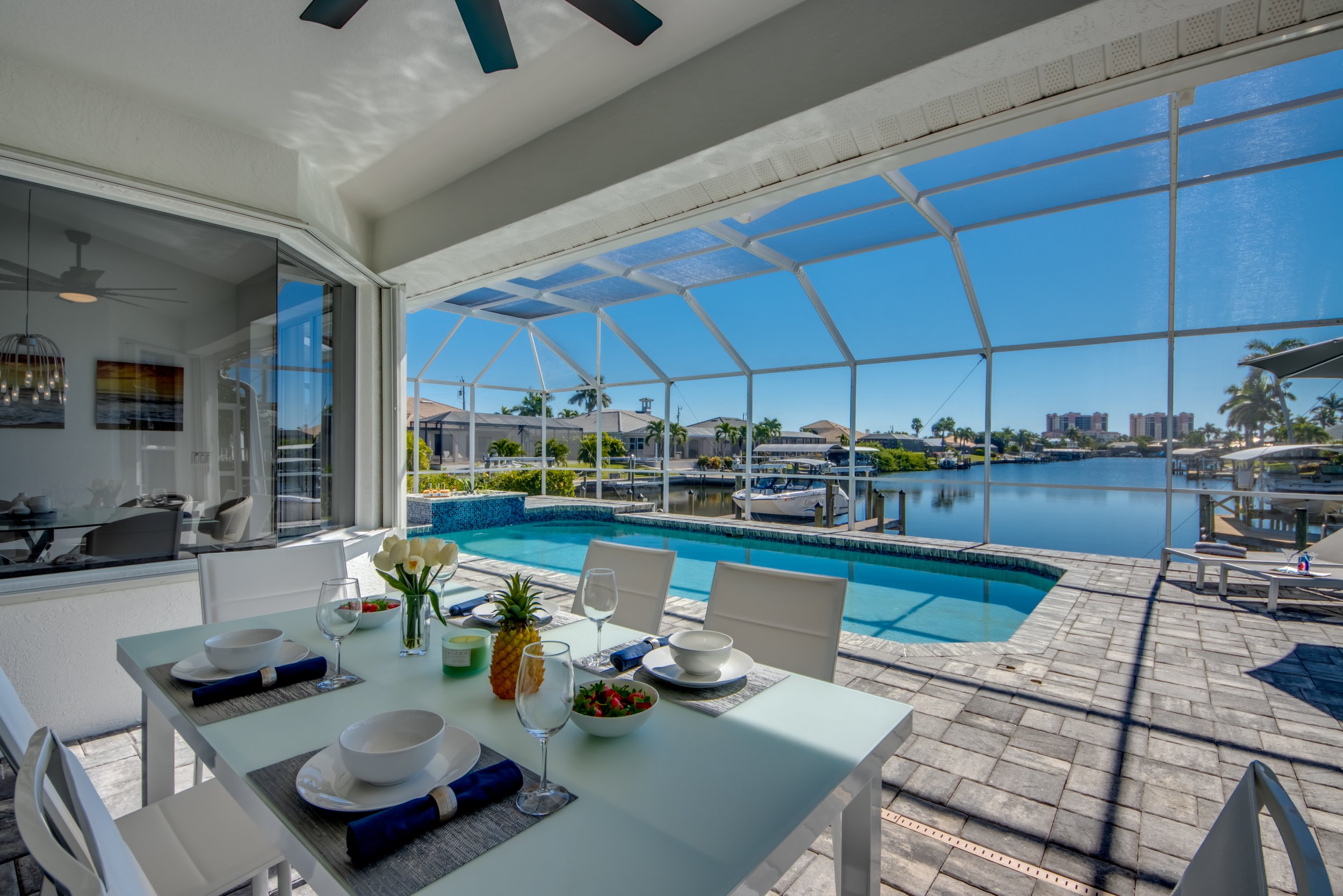 A dining table set with plates, bowls, glasses, and utensils, overlooking a screened-in patio area with a swimming pool and a view of a davenport canal with boats and waterfront houses under a clear blue sky.