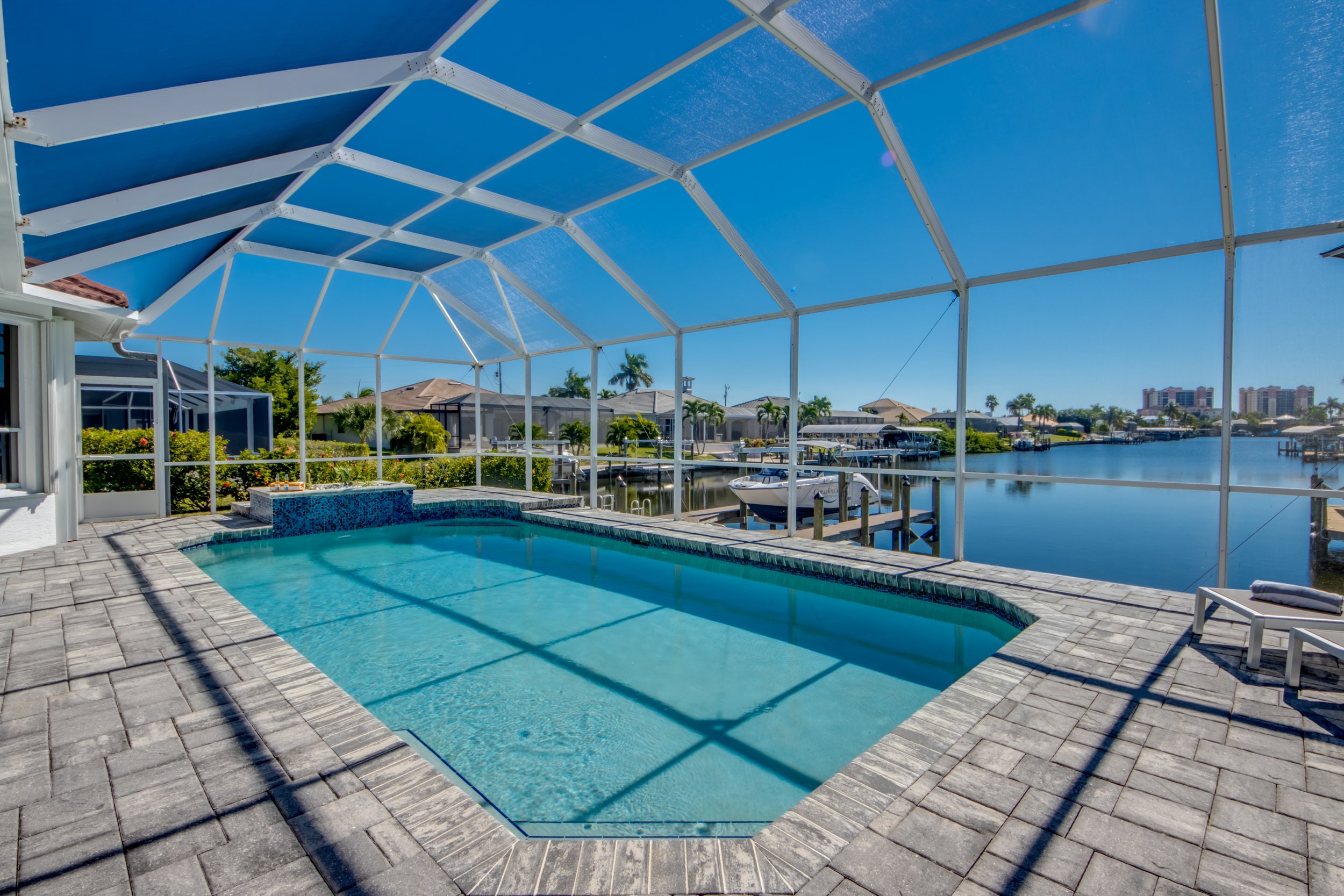 View of a screened-in backyard pool area overlooking a waterway with boats and residential houses, under clear blue skies.
