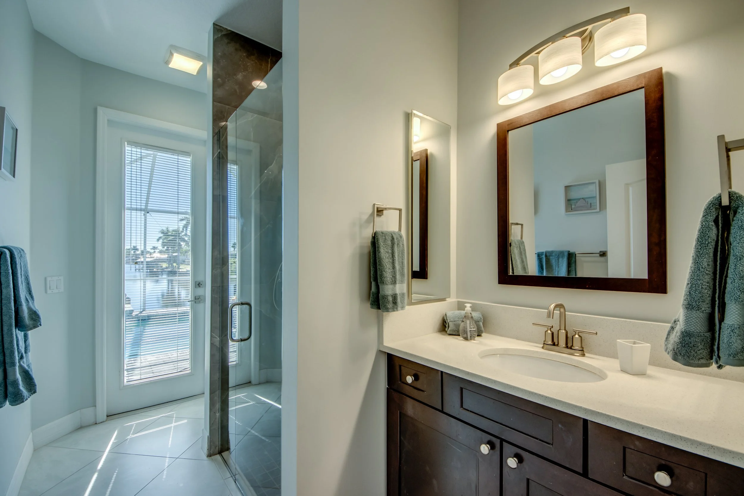 A bathroom with a white vanity, dark wood cabinets, a large mirror, and three modern light fixtures above the mirror. There is a towel rack with towels, a soap dispenser, and a linen tissue box on the countertop. To the left, a glass shower door and 