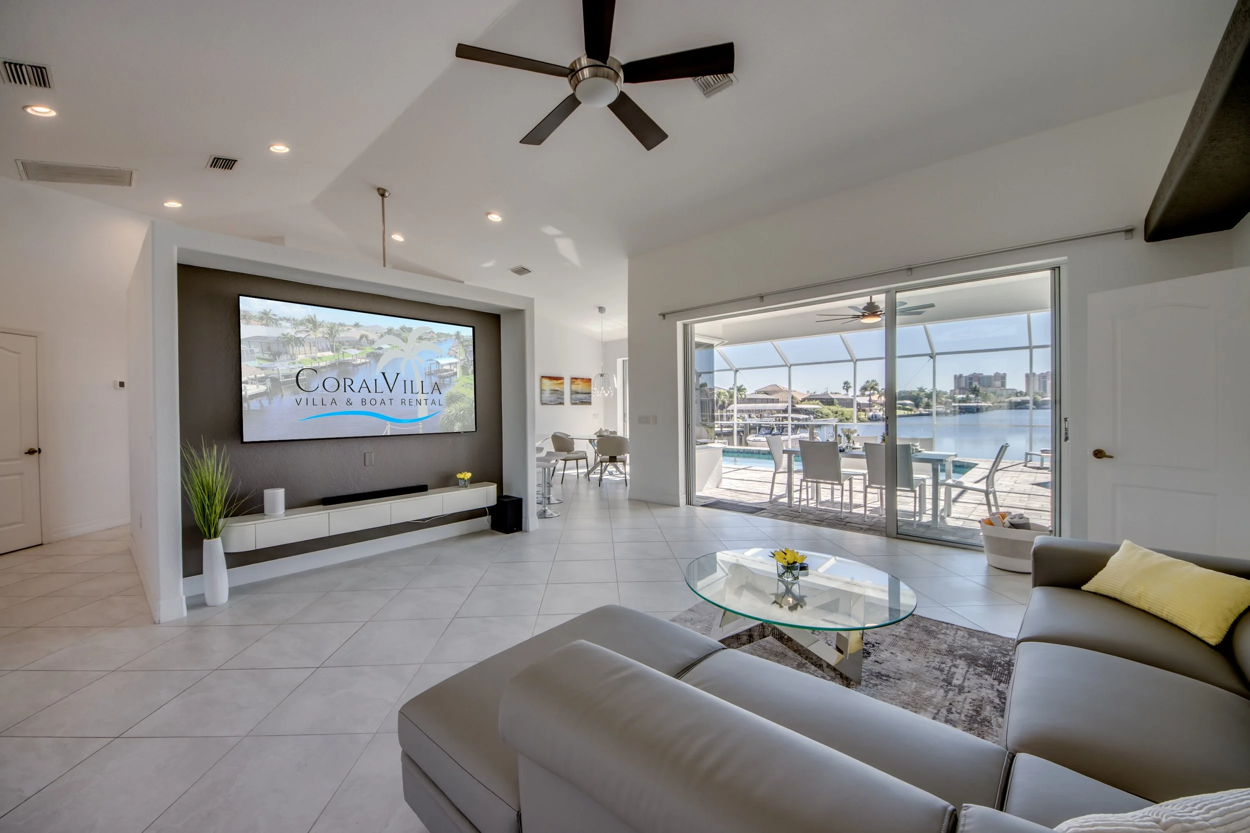 Modern living room with white sofa, glass coffee table, large sliding glass door, and a view of a pool and waterway outside.