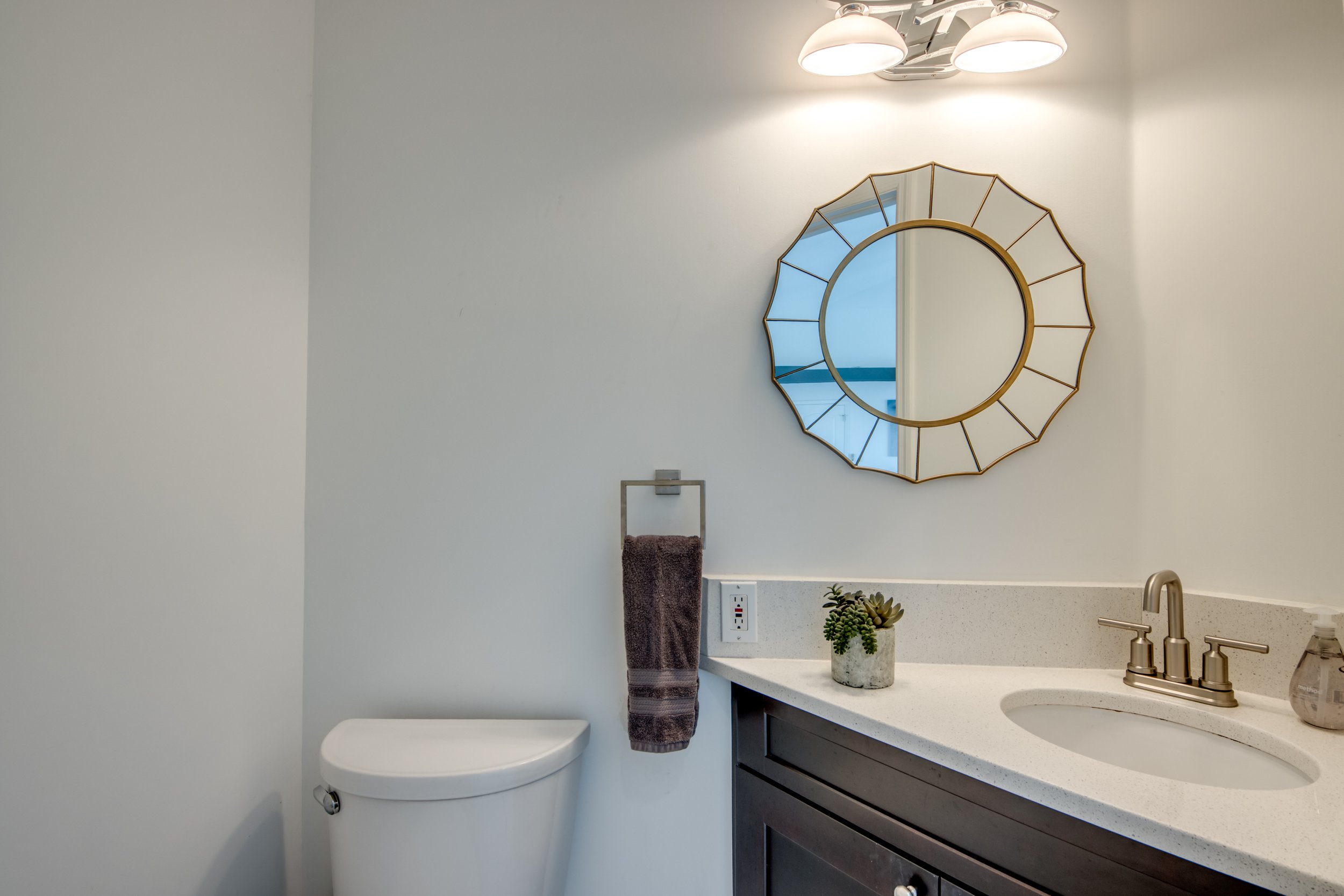 Bathroom with a round mirror on white wall, white countertop with sink, beige faucet, potted succulent, brown towel, electrical outlet, and a white toilet.