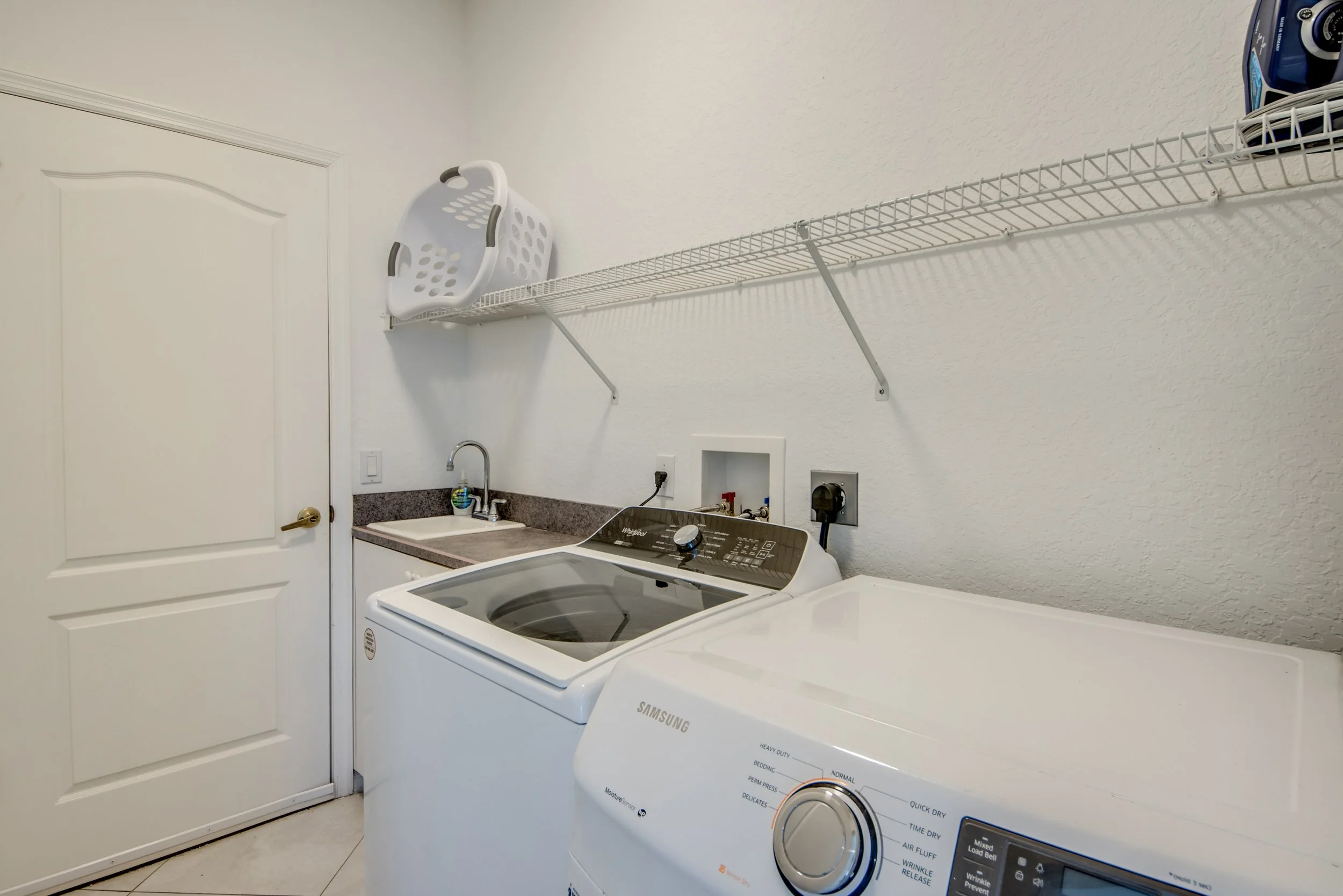 A laundry room with a white door, a white laundry basket hanging on the wall, a sink with a faucet, a countertop, and two white washing machines side by side, one of which is a Samsung model.