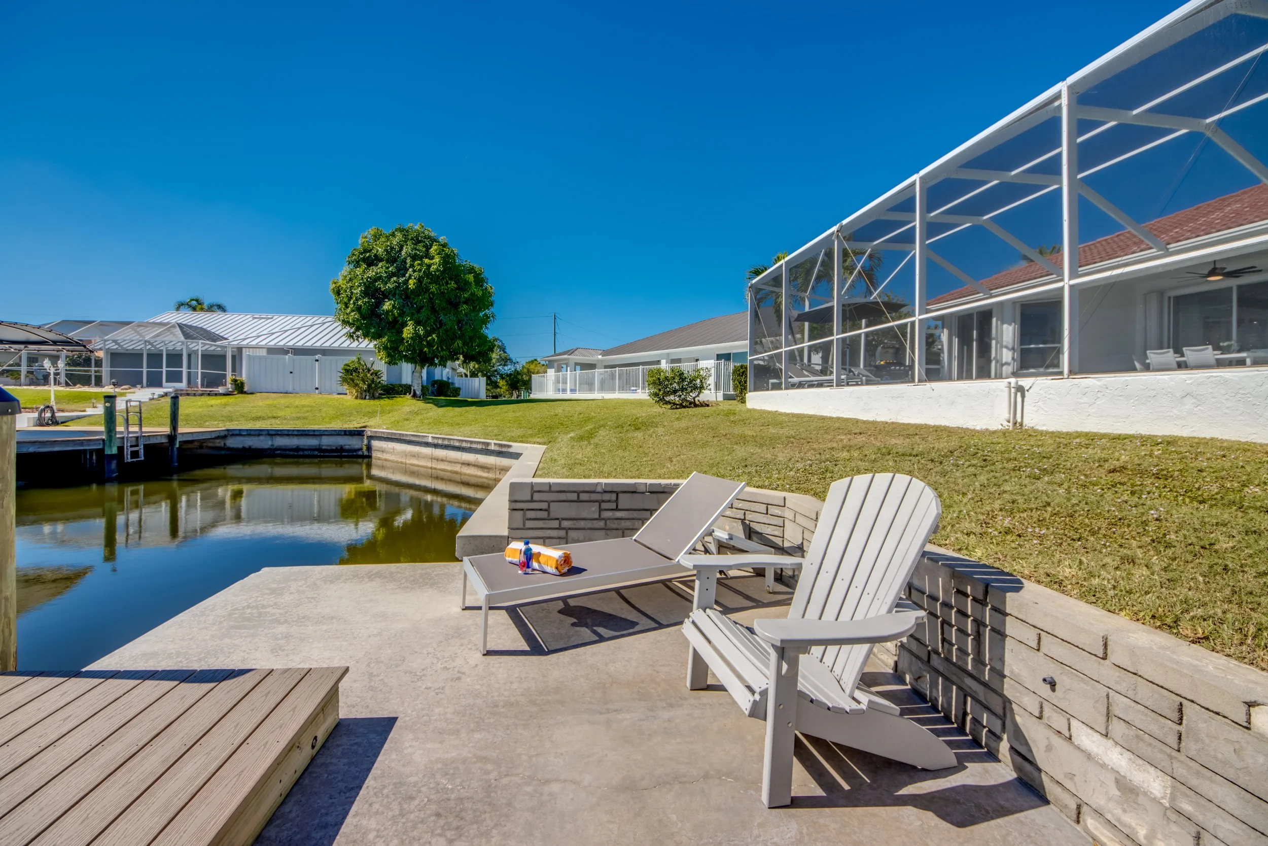Backyard patio with white Adirondack chairs, a small table with drinks, a swimming pool, and screened porch area, under clear blue sky.