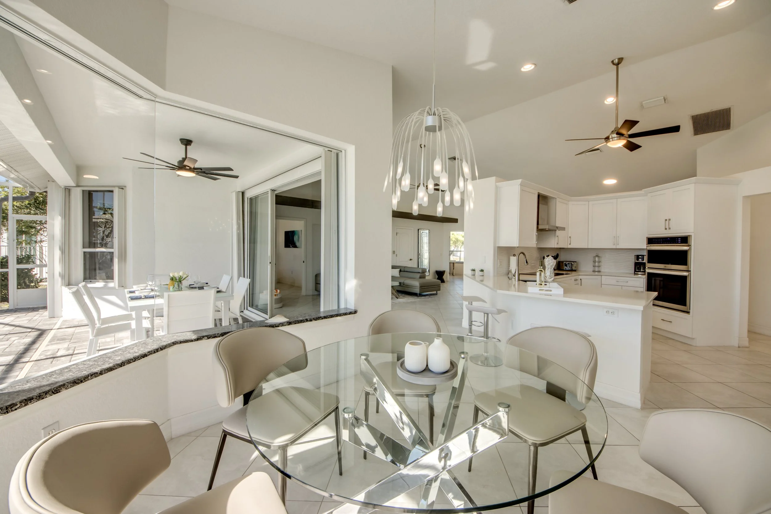 Bright, modern kitchen with white cabinets, a white island with bar stools, stainless steel appliances, and a unique chandelier hanging above the dining table. There is also a connected sunlit dining area with white chairs and tables, large sliding g