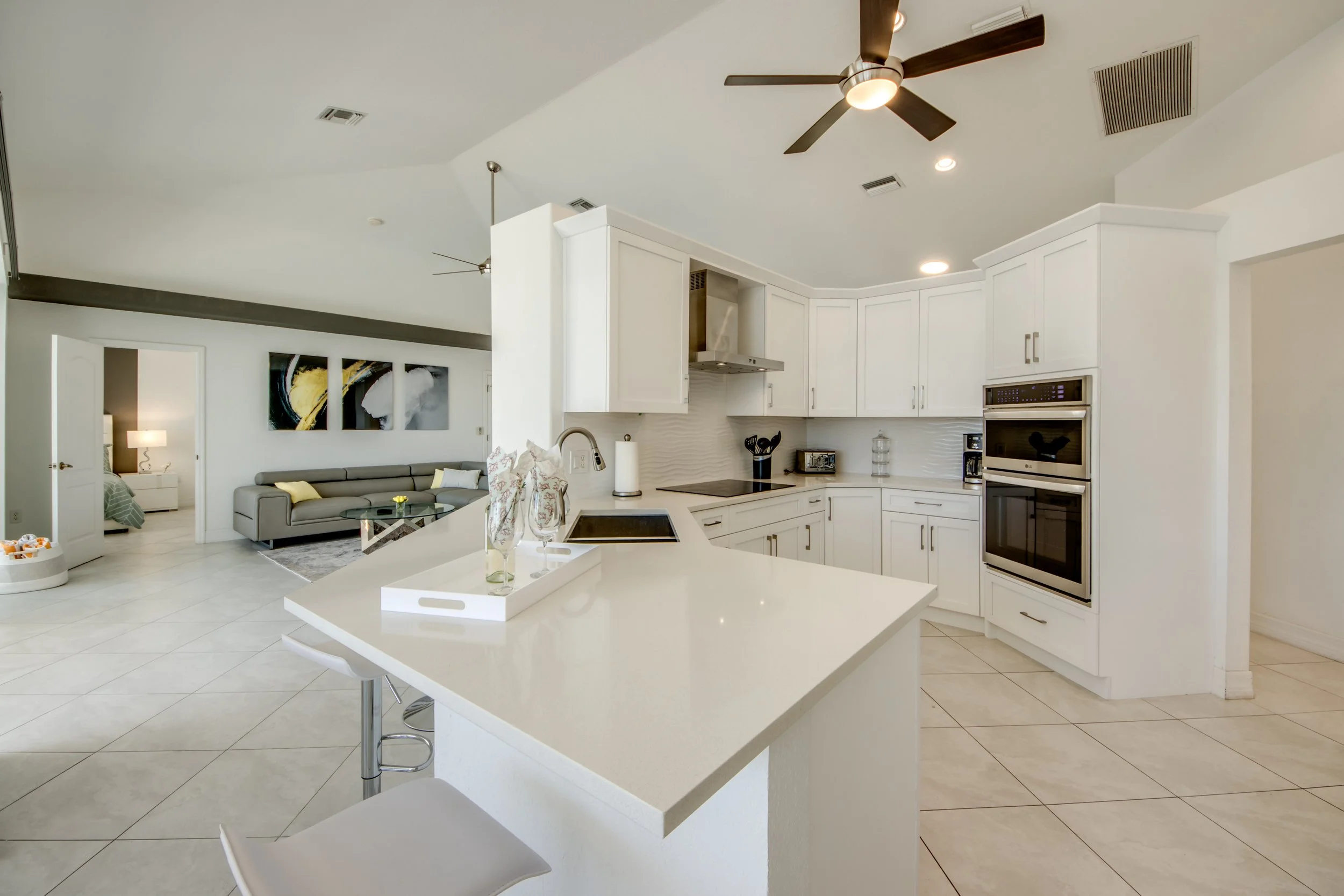 Modern kitchen with white cabinets, double built-in oven, and island with barstools. Open living area with gray sofa and abstract art on the wall.