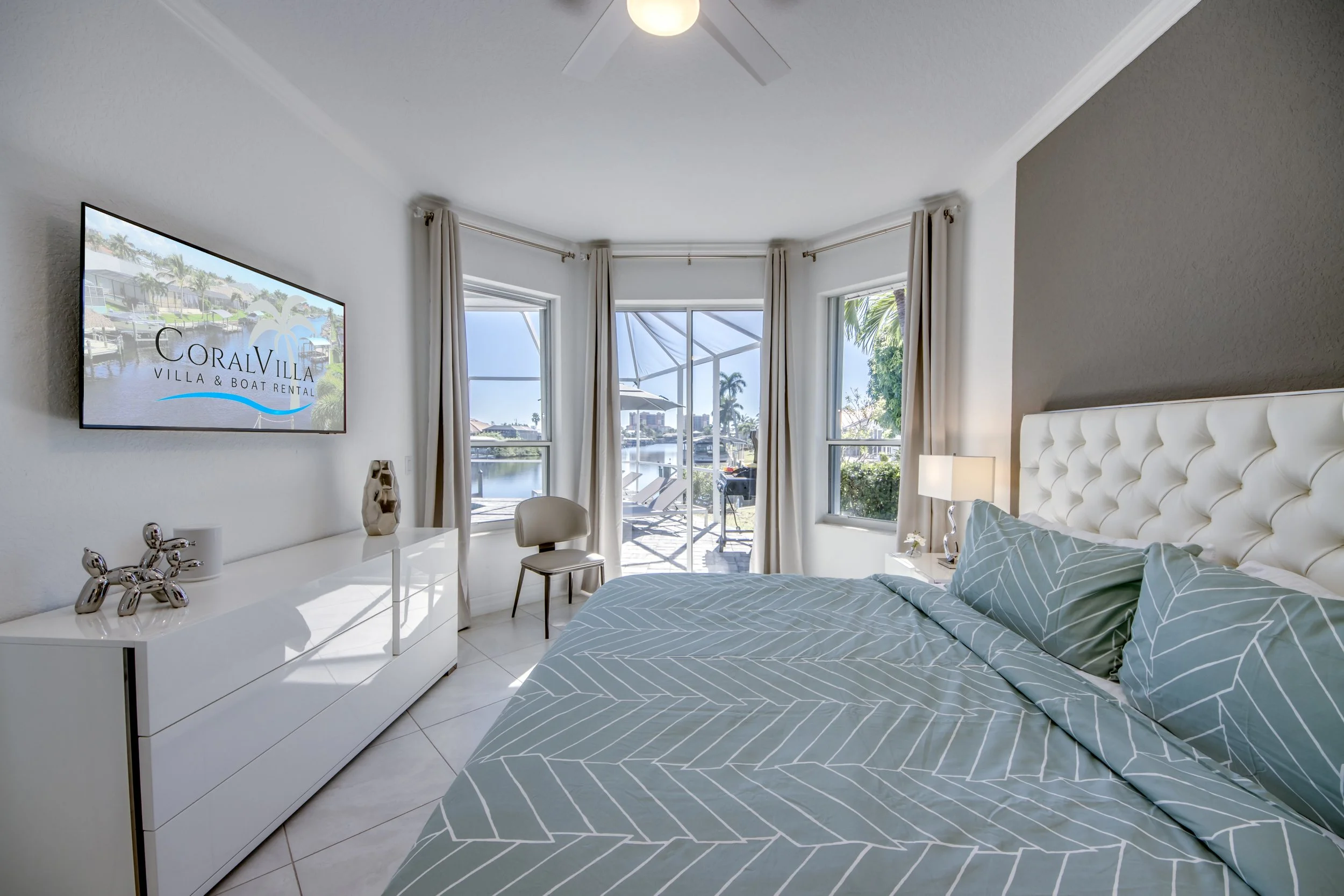 A bedroom with a large bed covered with a gray and white geometric patterned duvet and pillows, a white dresser with decorative items, and large windows with white curtains overlooking a patio and waterway with boats.