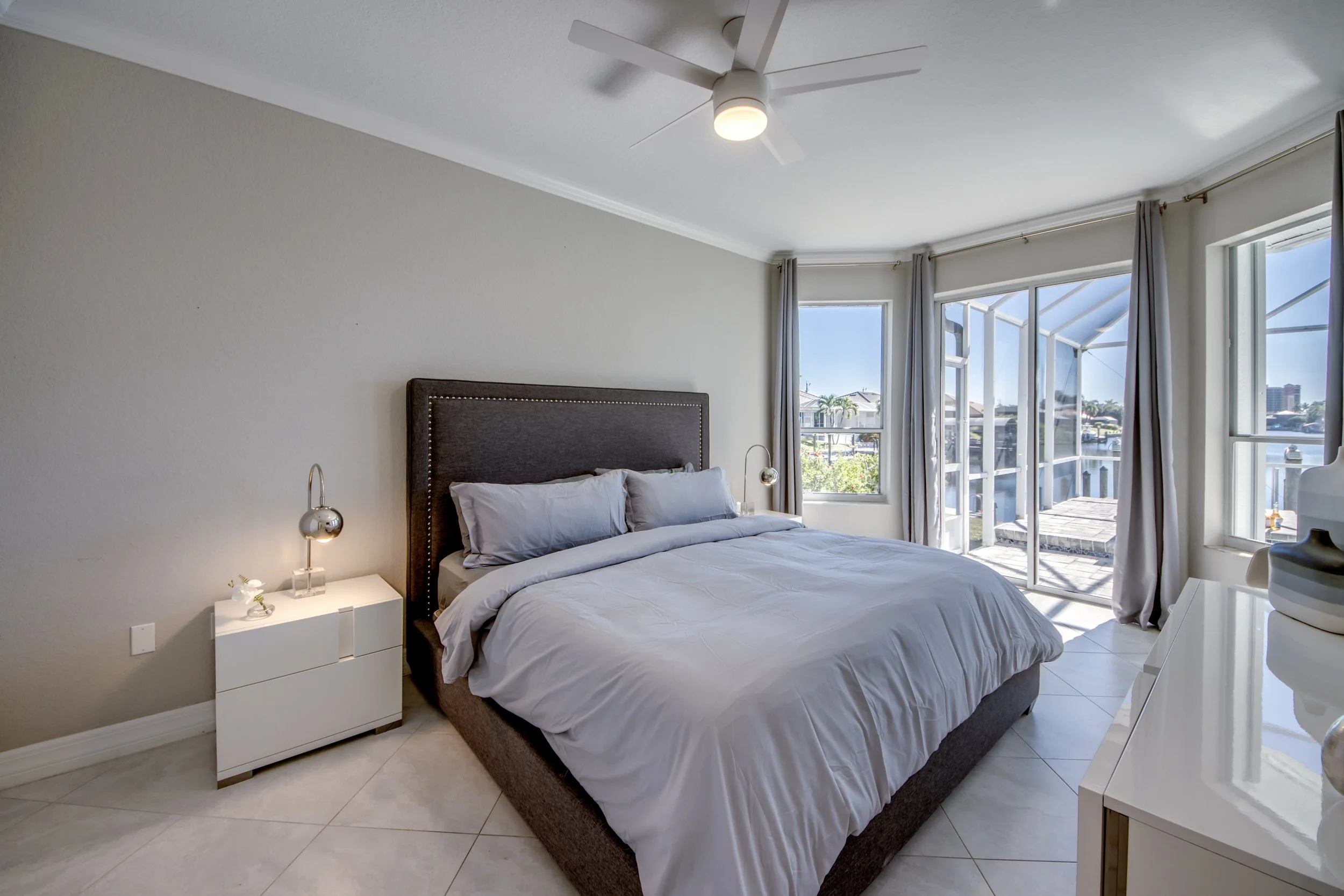 A bedroom with a bed, nightstand, and lamps, large windows with curtains and a view of neighboring houses and a balcony outside.