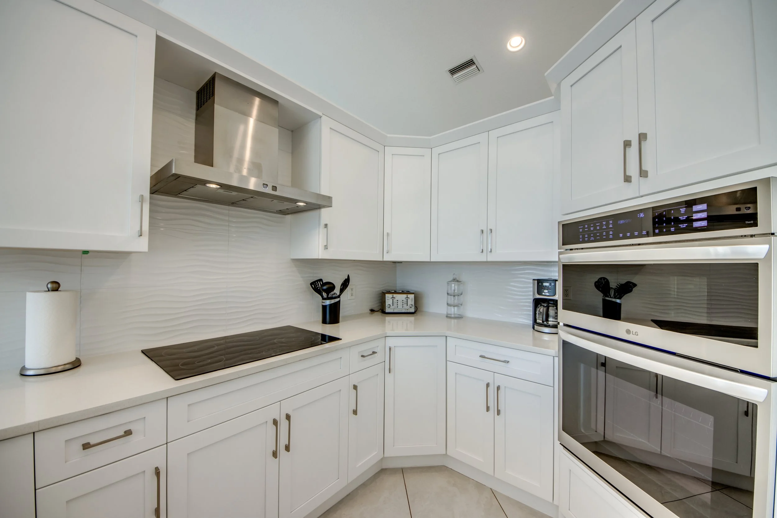 White kitchen with cabinets, black stovetop, stainless steel range hood, microwave, coffee maker, paper towel holder, and various kitchen utensils on the counter.