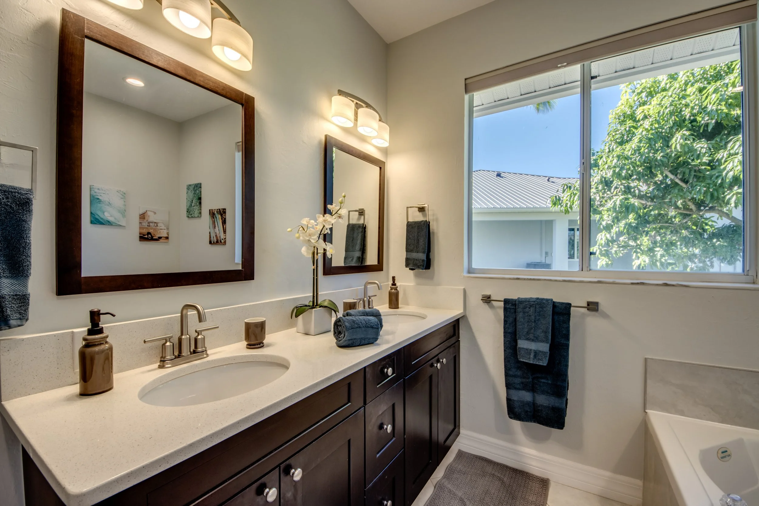 Bathroom with double mirror vanity, dark wood cabinets, white countertop, stainless steel fixtures, soap dispenser, potted orchid, towels, large window showing trees and rooftops, wall art, light fixture above mirrors, and a textured floor mat.