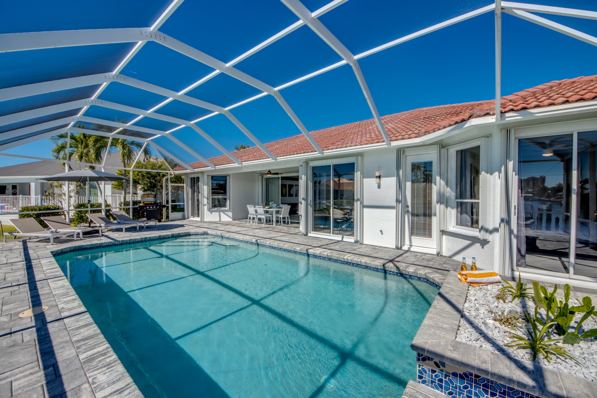 A backyard with a swimming pool, poolside chairs, and a patio table under a covered patio. The house has white exterior walls, a tiled roof, and large glass doors. An open outdoor summer setting with clear blue skies.