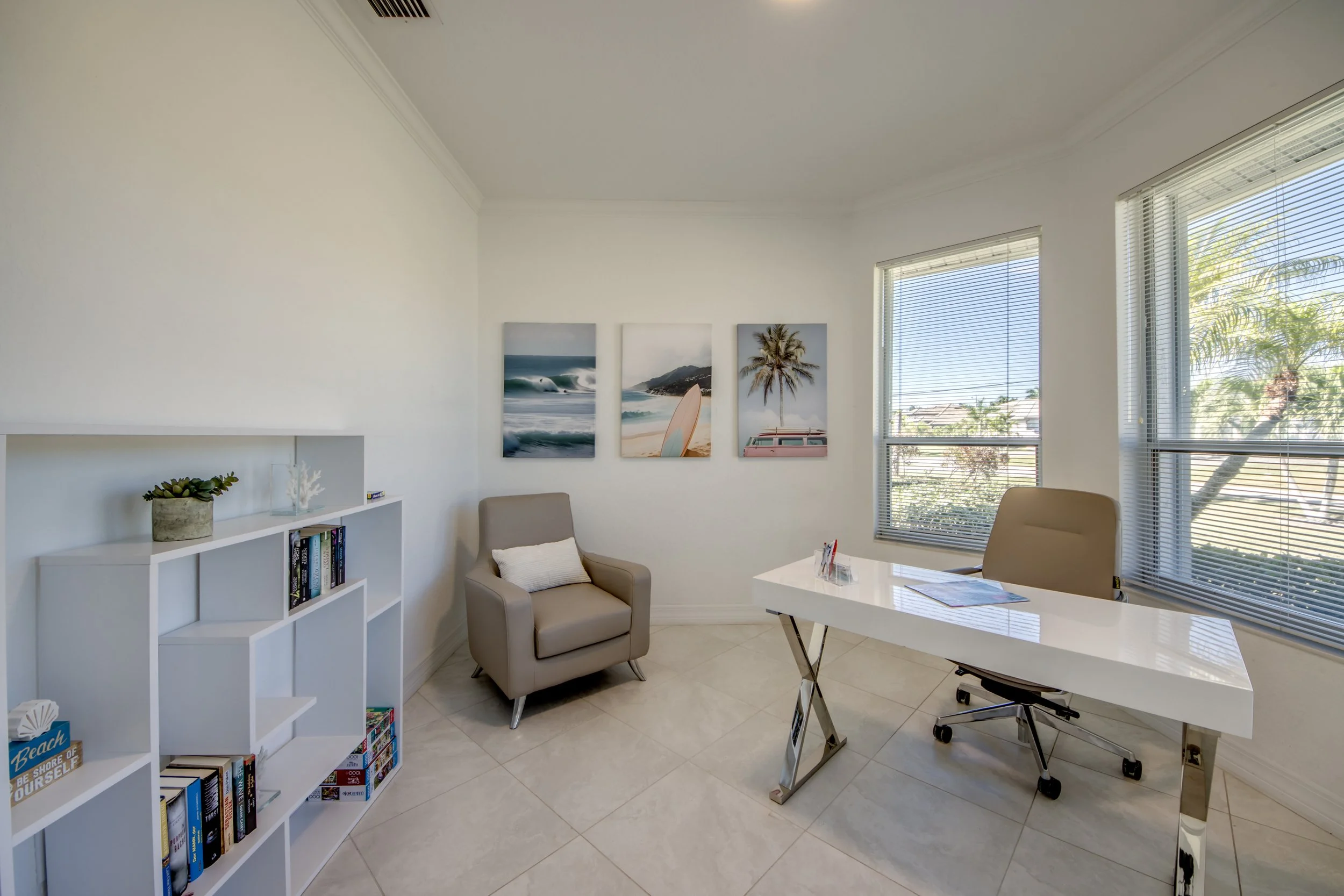 An office with large windows showing trees outside. Inside, there is a white desk with a chair, a beige armchair with a pillow, a white bookshelf with books and decorative items, and framed beach-themed artwork on the wall.