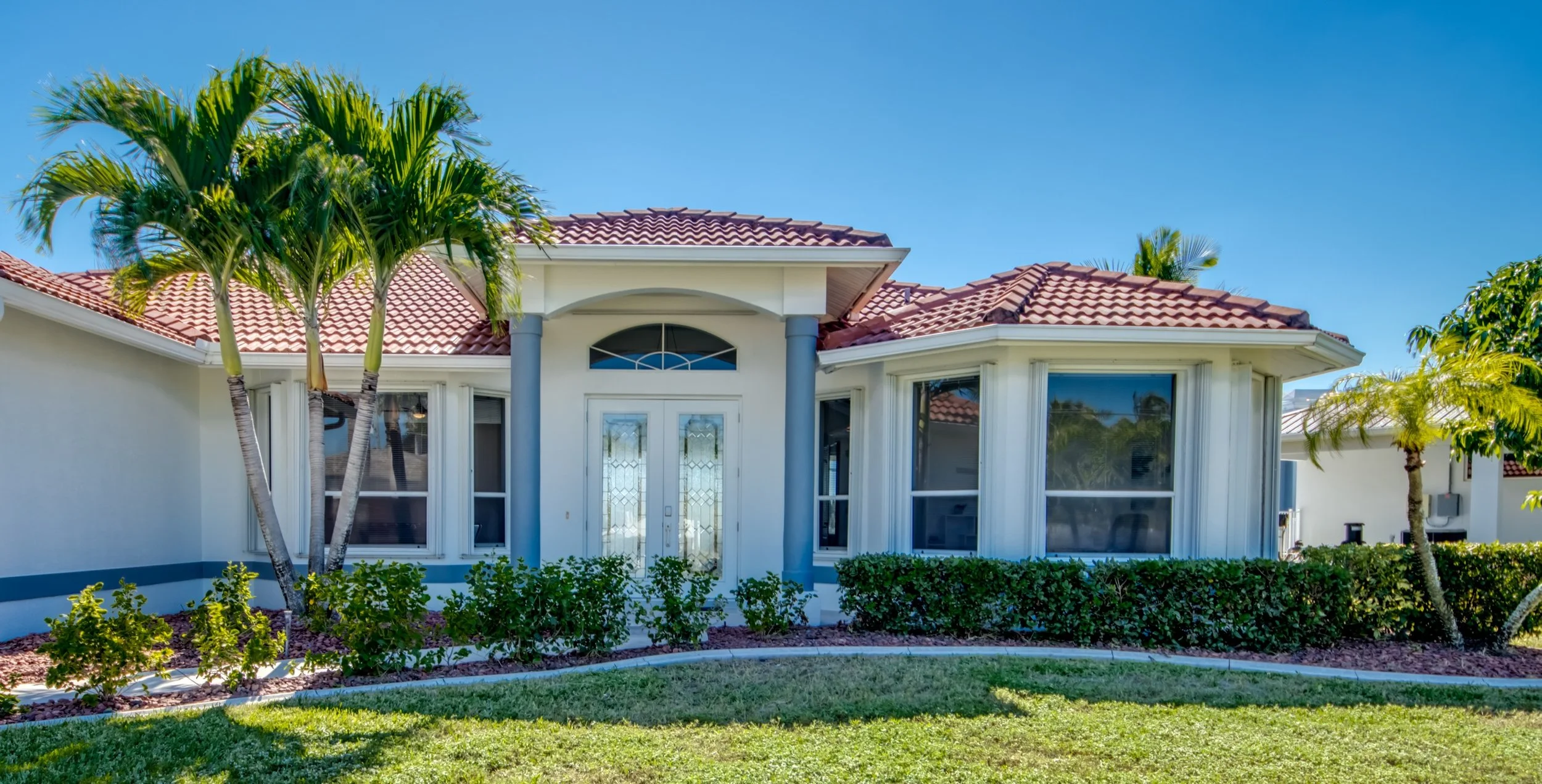 A house with a red tiled roof, white walls, and large windows, surrounded by palm trees and a well-maintained lawn under a clear blue sky.