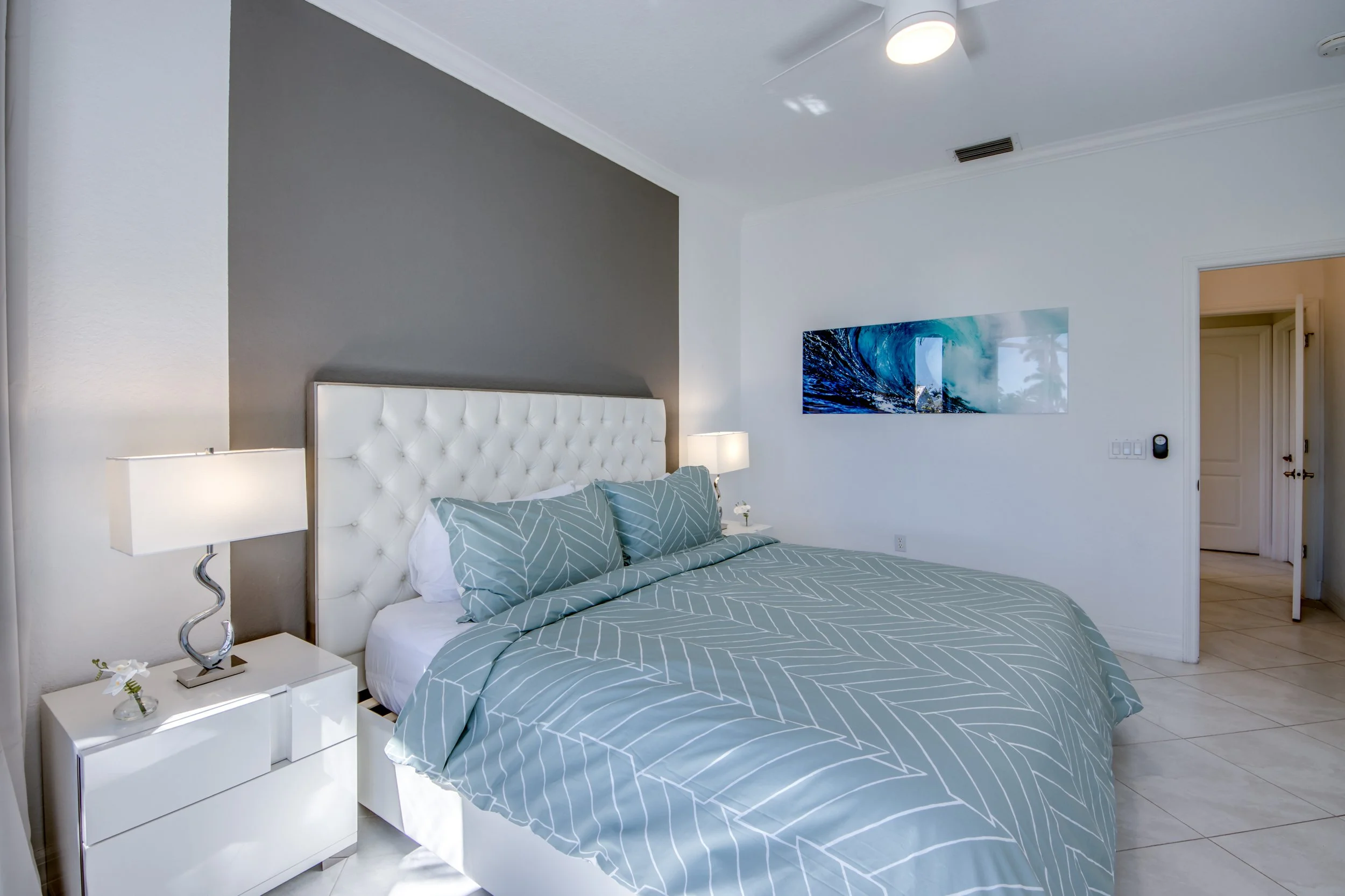 A bedroom with a white tufted headboard, two bedside lamps, a blue and white striped bedspread, and a modern abstract painting of ocean waves on the wall.