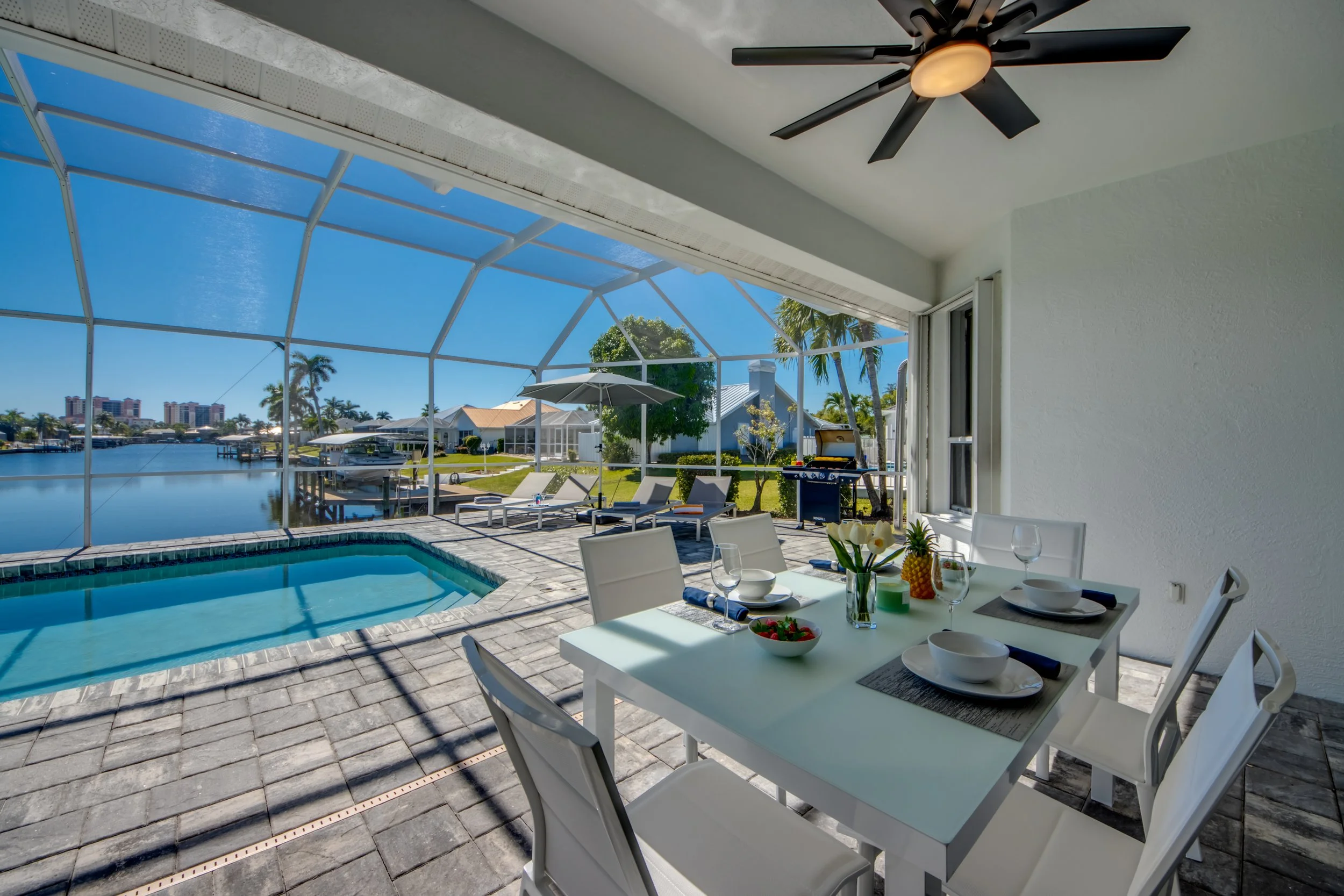 Patio area with pool and waterfront view, furnished with an outdoor dining table set with plates, glasses, and a bowl of strawberries, shaded by an umbrella, enclosed by a screened structure, with lounge chairs and a barbecue grill nearby, overlookin